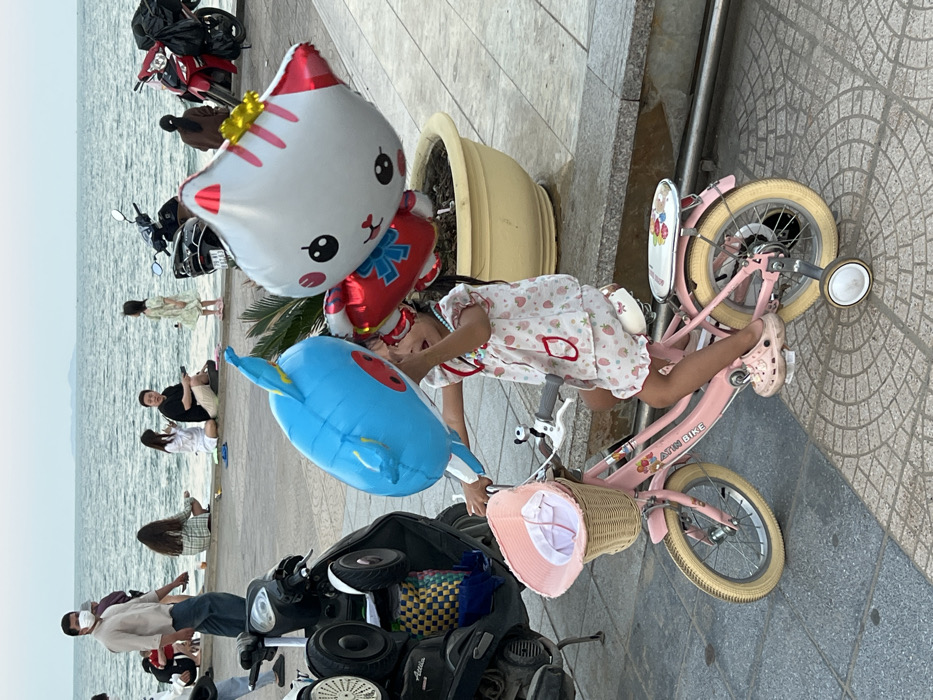 The image depicts a paved promenade in Nha Trang, Vietnam, adjacent to the ocean. In the foreground, a young girl, wearing a white two-piece outfit with strawberry patterns and pink clogs, is seated on a pink bicycle equipped with training wheels. She holds a cluster of balloons, prominently featuring a large blue whale and a white Hello Kitty head with a red bow, along with a smaller red balloon. The bicycle's front features a wicker basket containing a pink sun hat and a small white and black dog ornament on the handlebars. In the midground, several parked scooters are visible, one black and one grey, with a man in a light-colored shirt and face mask walking to the left. Along the stone barrier separating the promenade from the sea, multiple individuals are present: a group of three people are seated, one male examining an object, and another young girl stands near the water's edge. The ocean stretches into the background under a hazy sky, with distant land visible on the horizon. A large yellow potted plant stands on the right, partially obscuring a red motorcycle.