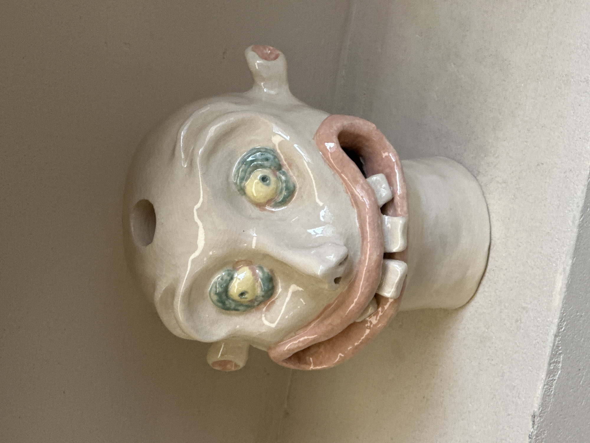 The image shows a ceramic sculpture of a head, off-white in color, with cartoonish features. It has two round, bulging eyes with green and yellow details. The mouth is depicted as a large, open cavity with exposed white teeth and pink flesh around the opening, resembling a gaping wound. The sculpture has a small protruding element at the top and bottom, possibly intended to represent ears or other facial features. There is a hole visible on the left side of the head. The head is attached to a cylindrical neck-like structure that is mounted to a wall. The sculpture is displayed in a corner space with light-colored walls. There is no indication in the image that it is in Paris, France.