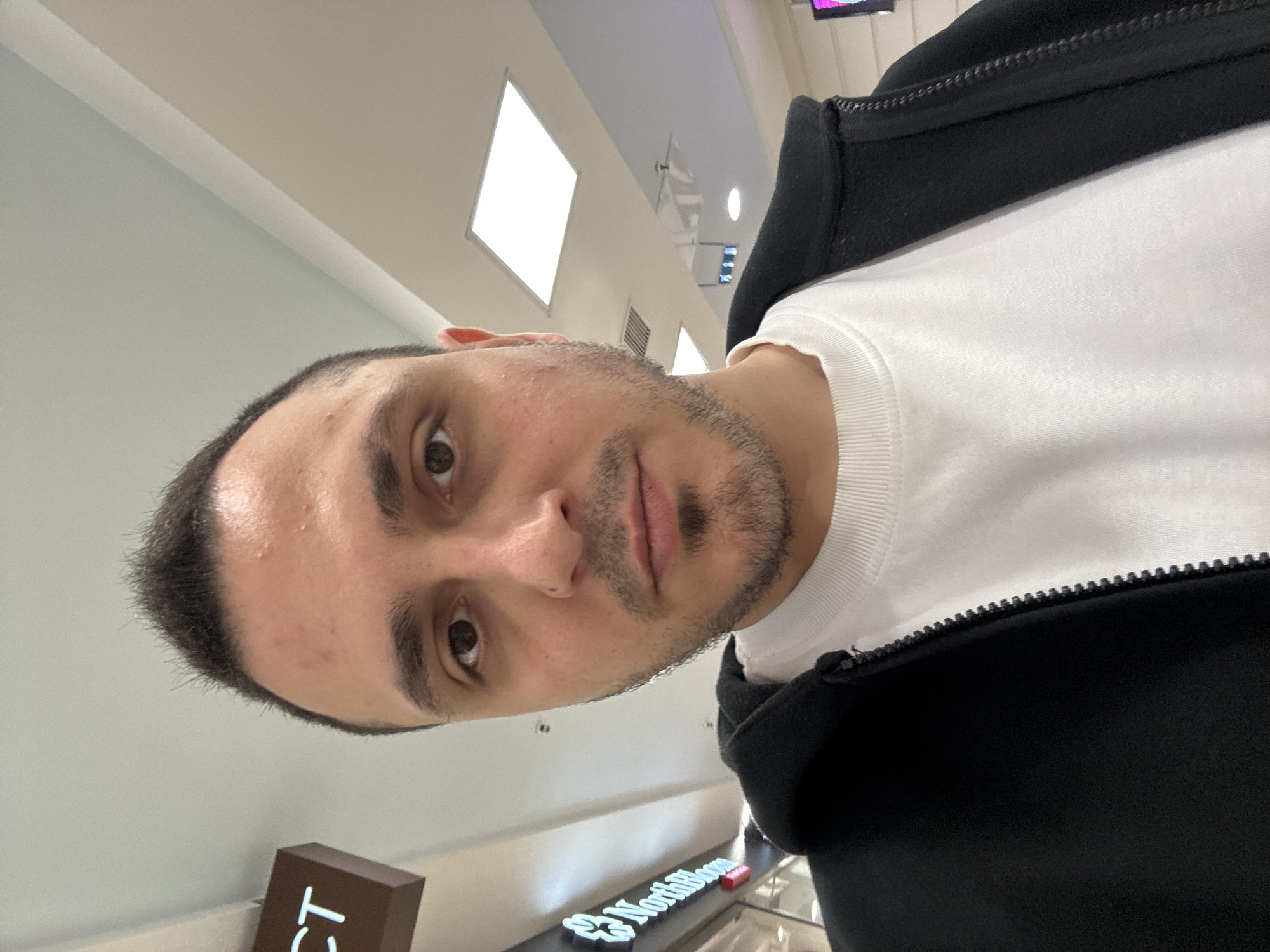 A man is taking a selfie in what appears to be a mall. He has short, dark, spiked hair and a goatee. He is wearing a black zip-up jacket over a white t-shirt. The background shows a ceiling with lights and a sign that reads "Northbloom".