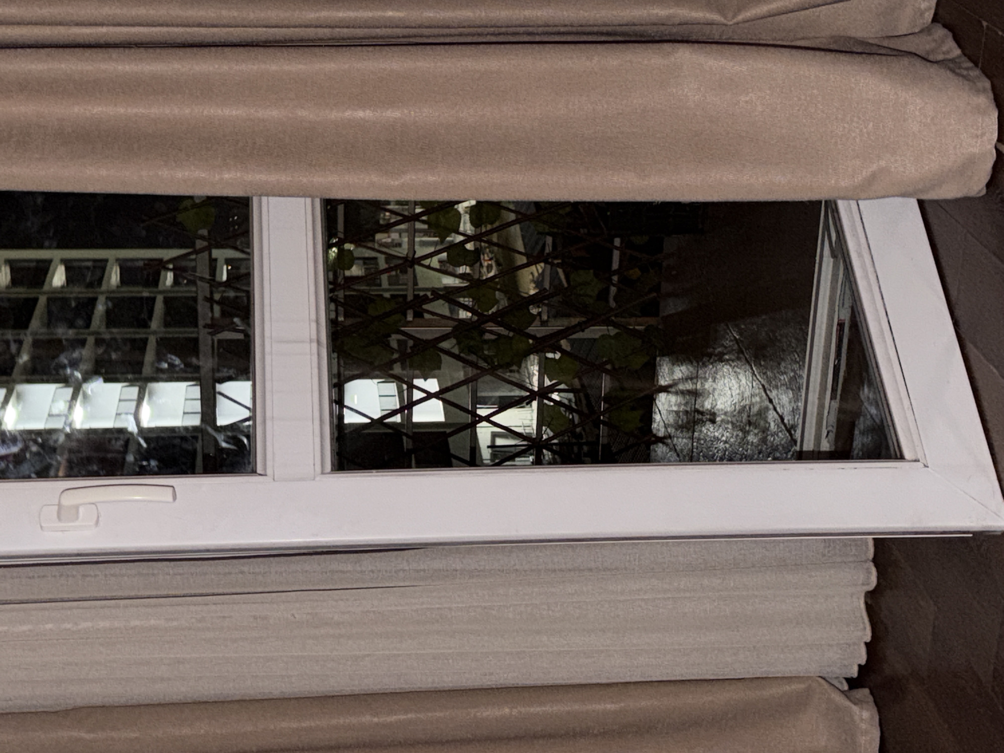 The image shows a partially open white framed window with a lattice screen inside. Light colored curtains frame the window on the left and right. A dark wood floor is visible at the bottom. Reflections in the glass show a building with multiple windows, some lit. The context provided indicates this is in Tbilisi, Georgia.