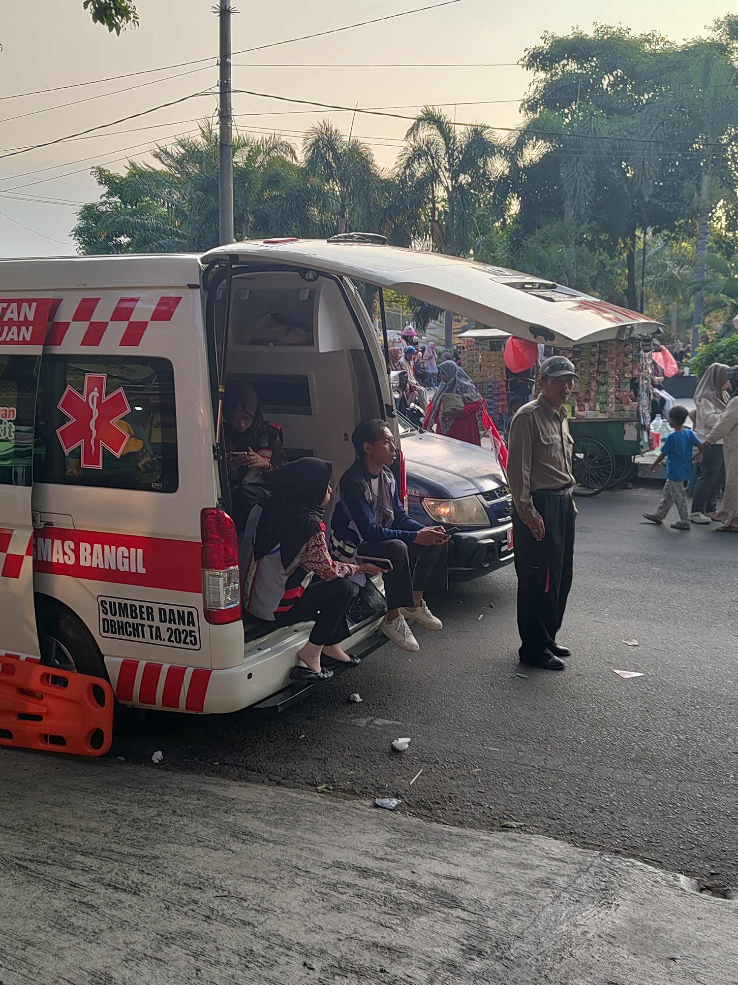 The image depicts an outdoor scene, likely in the city of Bangil, Indonesia, given the text on the ambulance. In the foreground, a white ambulance with its rear doors open is parked on an asphalt road. The ambulance has red and white checkered markings and an emergency medical symbol on its side, along with the text "MAS BANGIL" and "SUMBER DANA DBHCHT TA. 2025". An orange stretcher is attached to the side of the ambulance.
Several people are interacting with or near the ambulance. Inside the rear of the ambulance, two individuals, one appearing to be a woman wearing a hijab and dark clothing, and another person, are seated. A third person, a woman in a headscarf and patterned top, is also seated in the back of the ambulance, looking at her phone. Another individual, a young man wearing a dark jacket and white sneakers, is seated on the edge of the ambulance's open rear door, also looking at his phone.
Outside the ambulance, an older man in a brown long-sleeved shirt and dark pants stands facing away from the camera, appearing to observe the surroundings. To the right of the ambulance, a dark-colored car is parked. Further in the background, there are several food stalls, likely part of a street market or event. People are seen milling around these stalls and the surrounding area. Palm trees and other foliage are visible in the background, suggesting a warm climate. The sky is bright, indicating it is daytime. The ground is covered in asphalt and some concrete areas, with scattered litter visible.