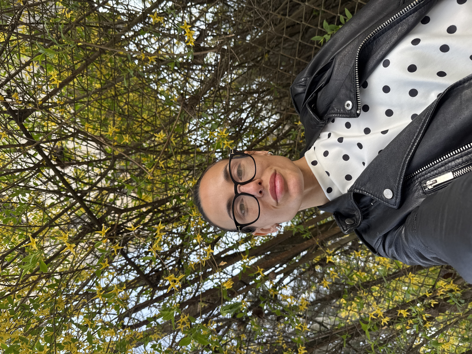 A woman with black-rimmed glasses and a black leather jacket over a white polka-dot blouse is standing outdoors in front of a bush with yellow flowers. The background appears to be a wire fence. The photo's location may be in the city of Pleven, Bulgaria.