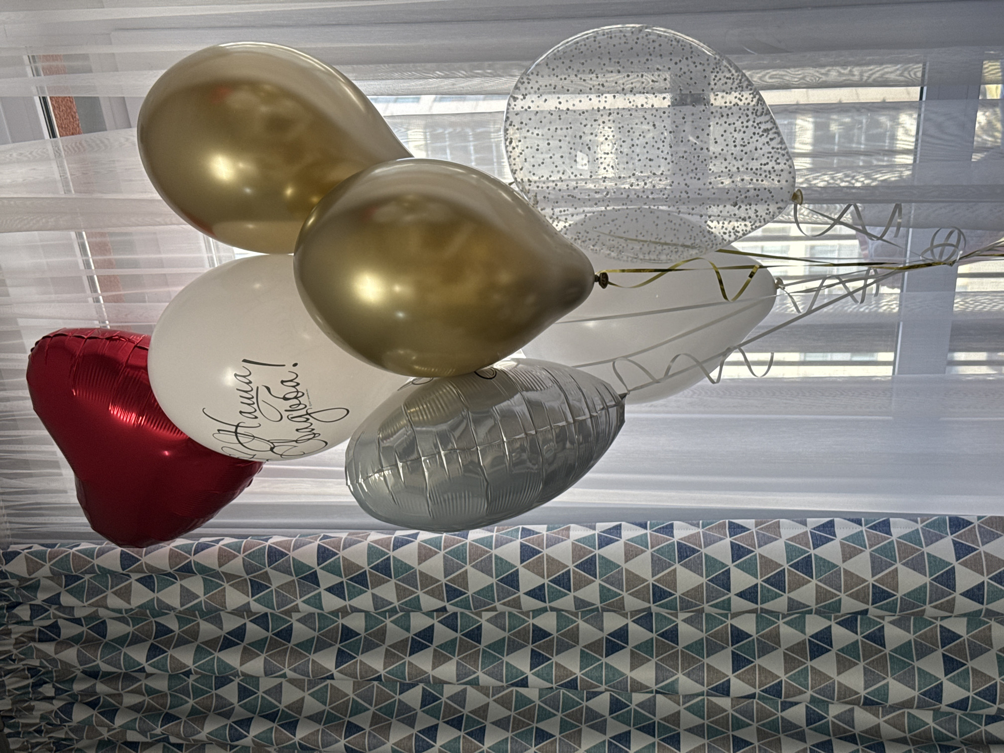 A cluster of helium balloons floats indoors, likely near a window, suggesting a celebratory occasion. The arrangement includes two metallic gold oval balloons, one clear balloon filled with small silver dots (possibly confetti), one metallic red heart-shaped balloon, one metallic silver oblong-shaped balloon, and one white oval balloon. Gold and silver curling ribbons are attached to some of the balloons.
The white balloon features black script text, which reads "Наша свадьба!" (Nasha svad'ba!), translating from Russian to "Our wedding!". This indicates the event is a wedding or wedding-related celebration.
In the background, a sheer white curtain or Venetian blinds partially cover a window, diffusing natural light into the room. Below the balloons, in the foreground, a patterned fabric can be seen, likely a roman blind or curtain. This fabric has a geometric design of interlocking triangles in shades of blue, grey, and white. The setting is a bright, festive indoor space, possibly in Izhevsk, Russia.