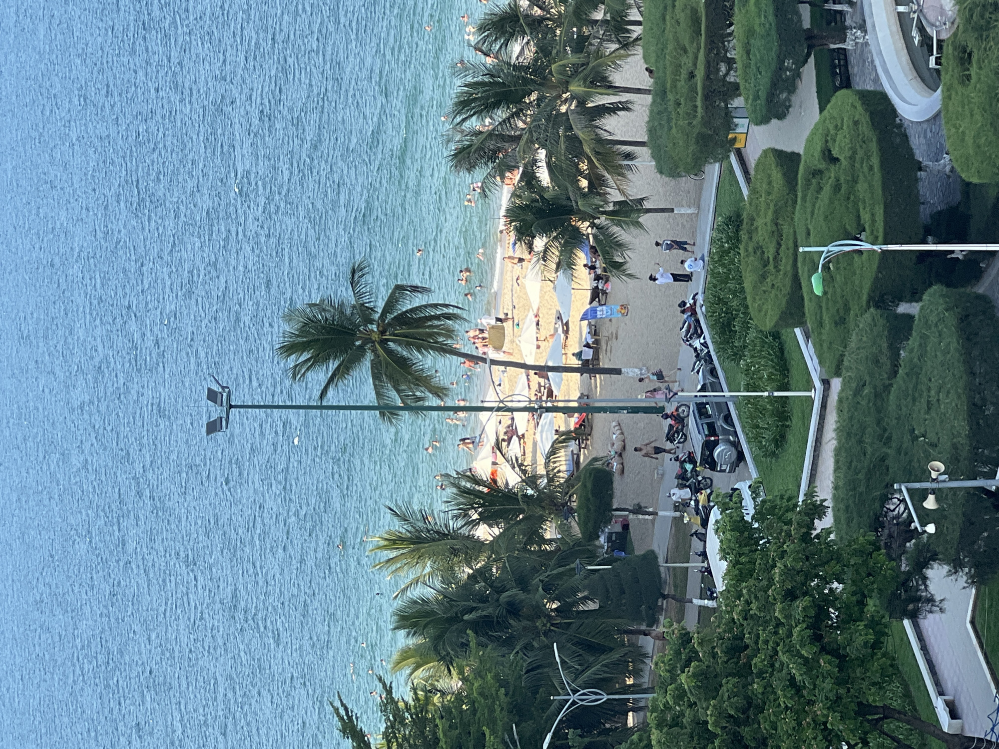 This image captures a vibrant scene at a beach in Nha Trang, Vietnam, likely taken from an elevated vantage point, possibly a hotel room or balcony. The cerulean ocean dominates the upper half of the frame, with numerous people visible swimming and enjoying the water.
The foreground transitions to a sandy beach dotted with beach umbrellas, lounge chairs, and scattered groups of people relaxing or walking. Lush green palm trees, a characteristic feature of Nha Trang's coastline, provide natural shade and add to the tropical ambiance.
Further inland, a well-maintained park area is visible, featuring neatly trimmed bushes and trees. A paved walkway runs through the park, and several motorcycles and a car are parked along the side, suggesting easy access to the beach area. A lamppost stands prominently on the beach, indicating nighttime lighting for the area. The overall atmosphere is one of a lively and popular tourist destination.