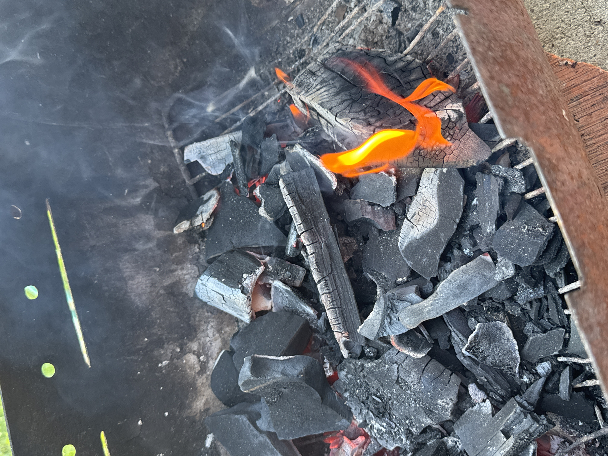 The image shows a close-up view of hot coals and burning wood inside a metal grill. Several pieces of charcoal are scattered across a metal grate, with some showing glowing red embers underneath. A few pieces of wood are actively burning, producing orange flames and wisps of smoke that rise upwards. The grill itself appears to be made of dark metal, with some rust visible on the edges. The background is blurred, but suggests an outdoor setting, possibly a yard or patio, with hints of green foliage and a concrete surface. There is no indication of any specific location within the city of Chaikovsky, Russia, nor are there any people or interactions depicted.