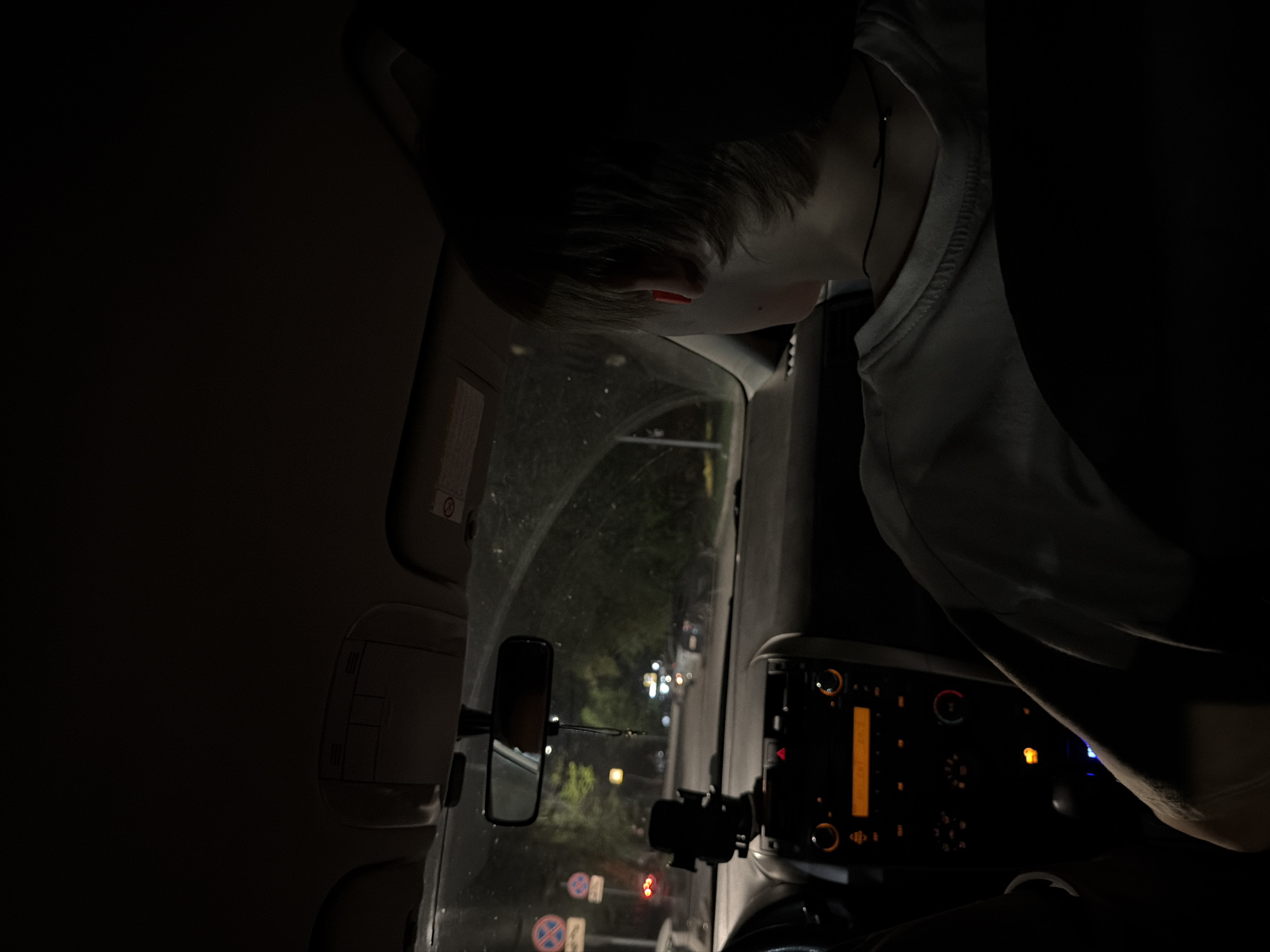 The image depicts the interior of a car at night, presumably in Izhevsk, Russia, based on street signs visible through the windshield. A person with short, dark hair is visible, leaning forward towards the dashboard. The car's dashboard is illuminated with various lights and displays, and a mobile phone holder is mounted on it. Through the windshield, streetlights and signs of the urban environment are visible. The overall light conditions are low, with most illumination coming from the car's interior lights and the streetlights outside.
