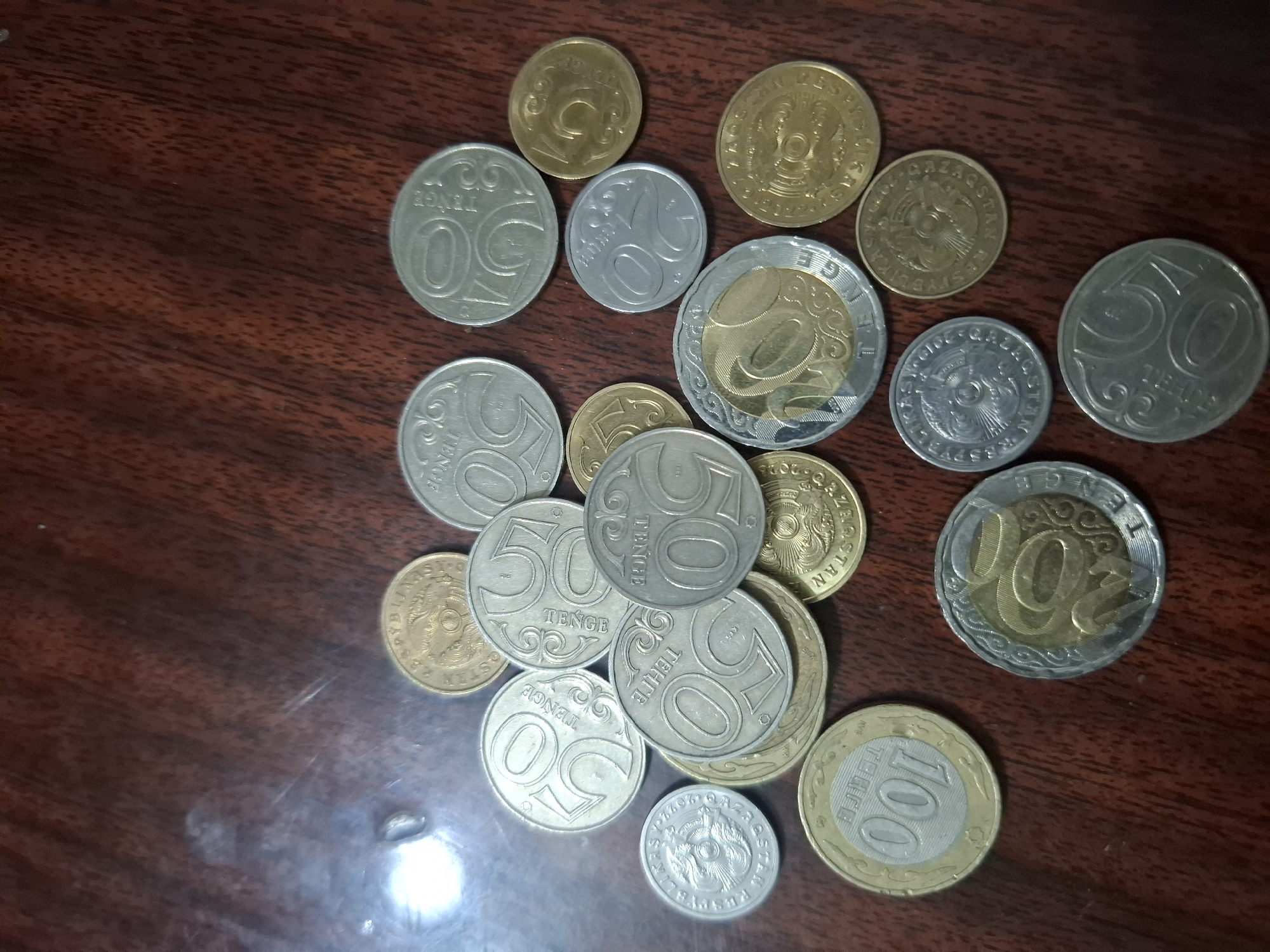 The image displays a collection of assorted coins scattered across a dark wooden surface, likely a table. The coins vary in size, color, and denomination, with some appearing to be silver and others gold or brass. Several coins clearly show the number "50" and the word "Tenge" (ТЕНГЕ), indicating they are Kazakhstani currency. One coin shows "100", and another appears to be "200". There are also coins with inscriptions and designs that suggest other origins or older mintages. The lighting in the image is uneven, with a bright reflection visible on the upper left side, suggesting a light source from that direction. There are no people or actions depicted, and the environment is simply the surface upon which the coins are placed. The specific location within Almaty, Kazakhstan, cannot be determined from the visual information provided.