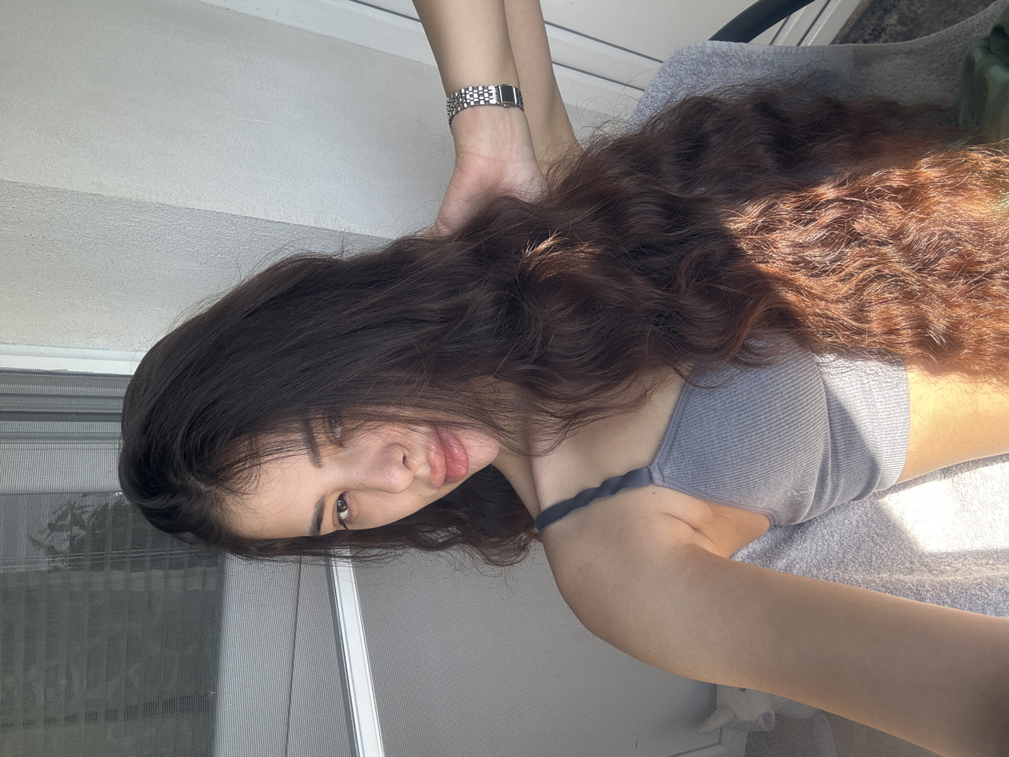 A woman with long, dark curly hair is lying down. She is wearing a gray top and a silver wristwatch. She appears to be next to a window. Without additional information, it's impossible to determine if the image was taken in the city of Plovdiv, Bulgaria.