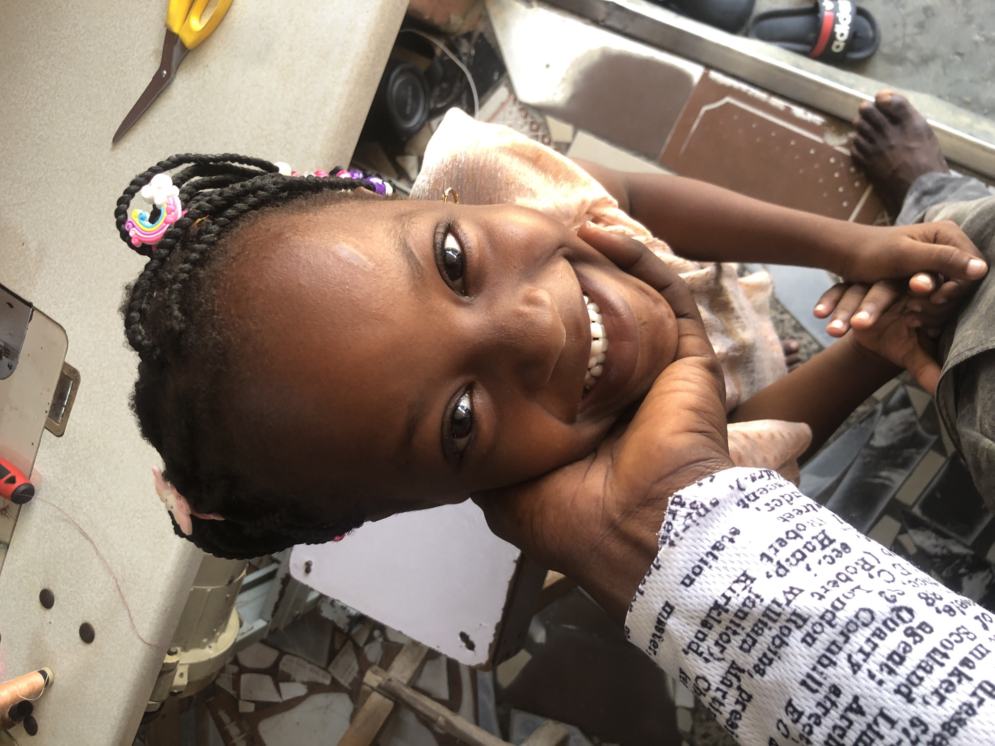 In a high-angle, medium shot, a young Black girl with braided hair is leaning her head against an adult's arm, looking up at the camera with a wide smile. She is wearing a light-colored garment, possibly a dress or a top, and her hair is adorned with colorful hair accessories, including one resembling a unicorn. The adult's arm, wrapped in a white bandage with black text, supports the girl's head.
The background suggests an indoor or semi-outdoor setting. To the left, a sewing machine with a white surface and some dark objects is visible, along with a piece of wood construction. To the right, a pair of yellow-handled scissors rests on the sewing machine's surface. Further to the right and in the background, a sliver of a black Adidas sandal with a red stripe is visible on the floor. The floor appears to be tiled with a pattern. The overall environment seems to be a workshop or a home with crafting materials.
The image appears to be taken in Ado, Nigeria, given the context. The lighting is bright, casting some shadows, and the focus is sharpest on the girl's face.
