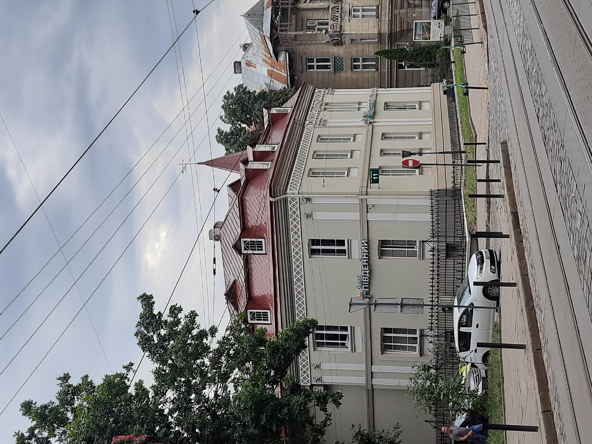 This is a street-level, eye-level, medium shot of a street scene in Lviv, Ukraine. The dominant structure is a light-colored, two-story building with a red-tiled, mansard roof. The building has white-trimmed windows and decorative white trim below the roofline. A sign on the building reads "Банк Південний" (Bank Pivdennyi). To the right of this building is a taller, older building with a weathered, rusty roof and ornate architectural details.
In the foreground, tram tracks run along a cobblestone street. A sidewalk separates the street from the buildings. Several cars are parked on the sidewalk area in front of the light-colored building, including a white hatchback. A person in a grey t-shirt is walking on the sidewalk to the left of the cars. Two green electric scooters are parked on the sidewalk area to the right.
The sky is overcast with grey clouds. Overhead, numerous electrical wires are visible, crisscrossing the frame. A street sign with the number "11" is mounted on the light-colored building. A red and white "no entry" sign is also visible on a lamppost in front of the building. A tree with green foliage is situated to the left of the light-colored building.