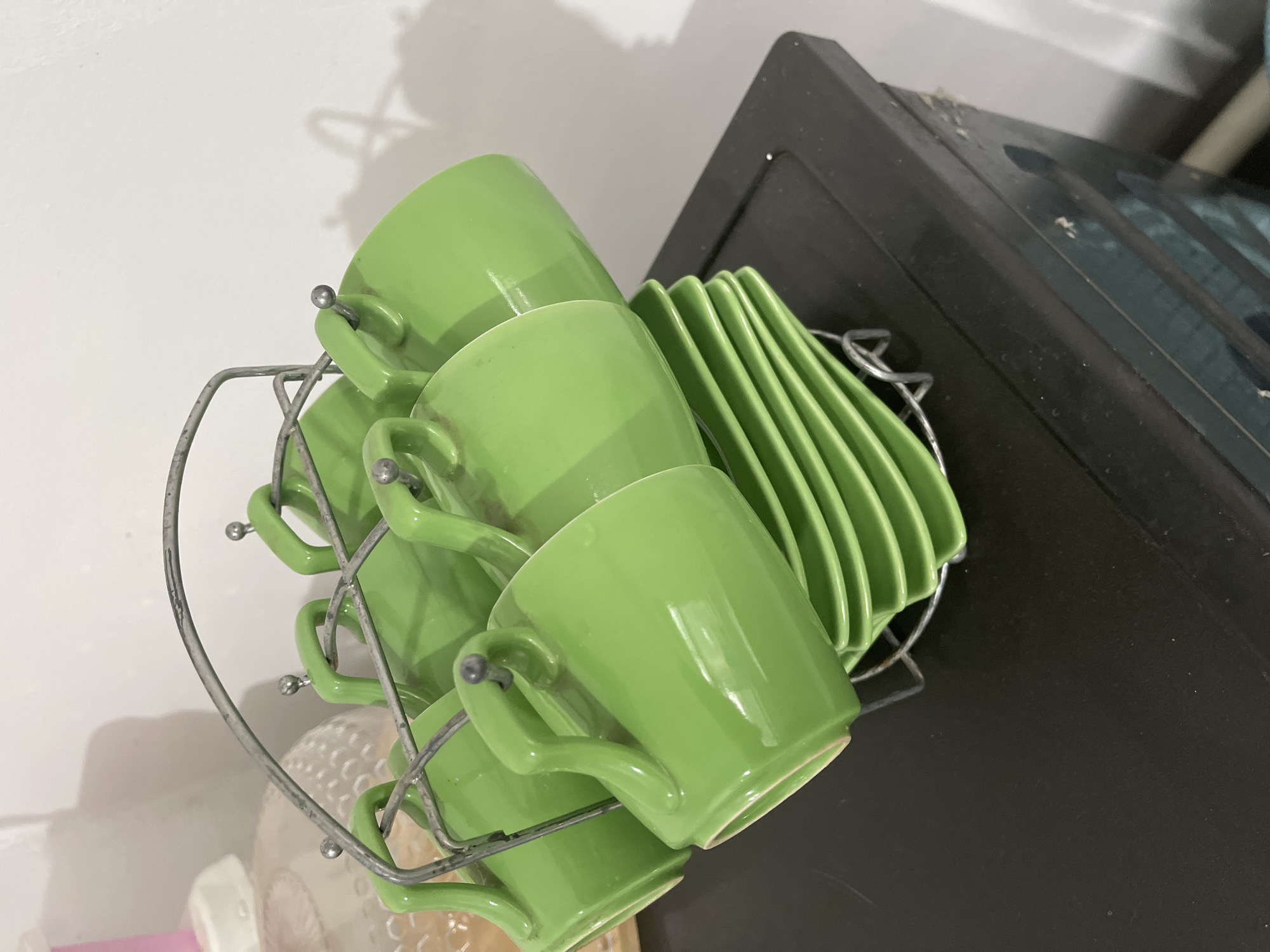A set of six bright green ceramic teacups and saucers is displayed on a metal rack. The rack is positioned on top of a dark brown cabinet, which appears to be made of particleboard with a laminate finish. The background is a plain white wall, showing a faint shadow of the teacup rack. To the left of the rack, a portion of a large, clear glass jar with a textured surface and a pink lid is visible. The overall scene suggests a domestic setting, possibly a kitchen or dining area. There are no people present in the image. The image does not provide any specific information about the location in Bishkek City, Kyrgyzstan.