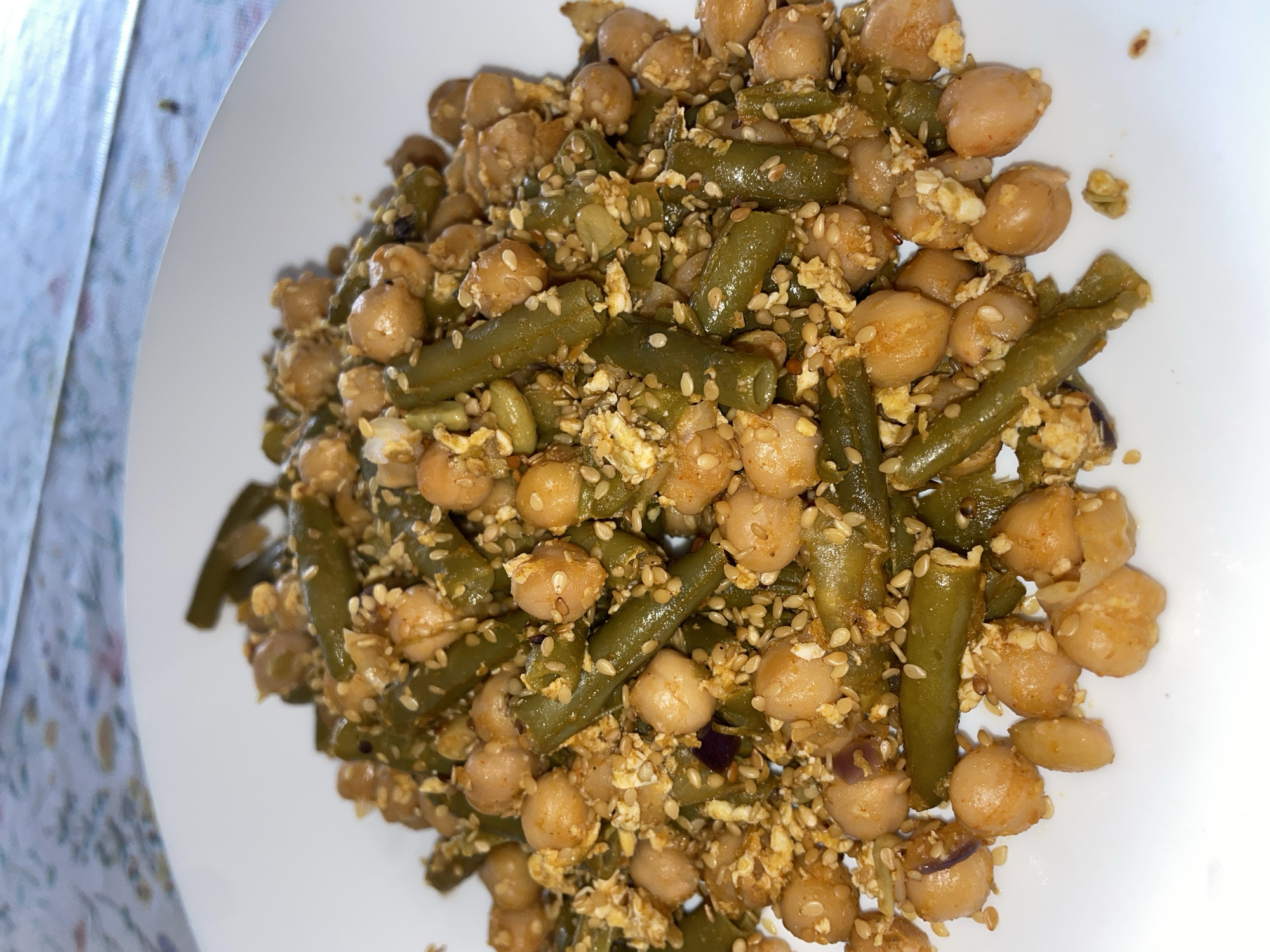 A top-down, close-up shot depicts a white plate filled with a mixture of cooked green beans and chickpeas, sprinkled with sesame seeds and what appears to be scrambled eggs. The food is piled in the center of the plate, with visible white edges suggesting a dining setting. The background shows a white and lightly patterned cloth, possibly a tablecloth or napkin. The scene likely occurs indoors, based on the controlled lighting. There's no evidence to suggest the specific location within the city of Senden, Germany; the setting is limited to a domestic or restaurant environment.