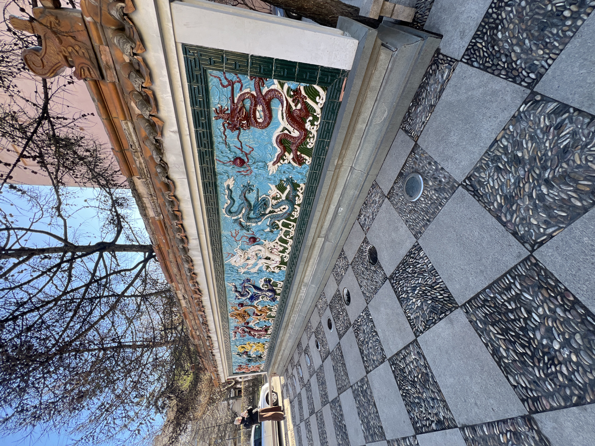 The vertically oriented image displays a detailed architectural feature alongside a patterned ground, beneath a clear blue sky. A long, ornate wall with a traditional East Asian roofline runs across the upper section. The roof features curved eaves, intricate brown wooden supports, and weathered red-orange tiles. Below this, a vibrant ceramic tile frieze dominates, showcasing multiple dragons in dynamic poses amidst stylized waves and clouds, often with a flaming pearl. These dragons are rendered in various colors, including red, blue, white, and gold, set against a light blue and white background and framed by a dark green geometric border.
The ground is paved in a distinctive checkerboard pattern. Alternate squares are made of smooth grey paving stones, while the others are filled with small, dark, varied-colored pebbles arranged in a mosaic. Several round, metallic ground lights are embedded within these pebble-filled squares.
To the left, bare tree branches, indicative of winter or early spring, are silhouetted against the bright, cloudless blue sky. A person, dressed in a dark jacket and light trousers, is partially visible near the bottom left, standing next to a white car parked adjacent to the wall.
The scene unfolds during clear, sunny daytime. The prominent Chinese architectural style and dragon motifs strongly suggest a cultural installation within a broader setting. Given the context of Saint Petersburg, Russia, this likely depicts a themed area, such as the Garden of Friendship on Liteyny Prospekt, which features similar traditional Chinese elements. No specific activity or visible text is apparent.