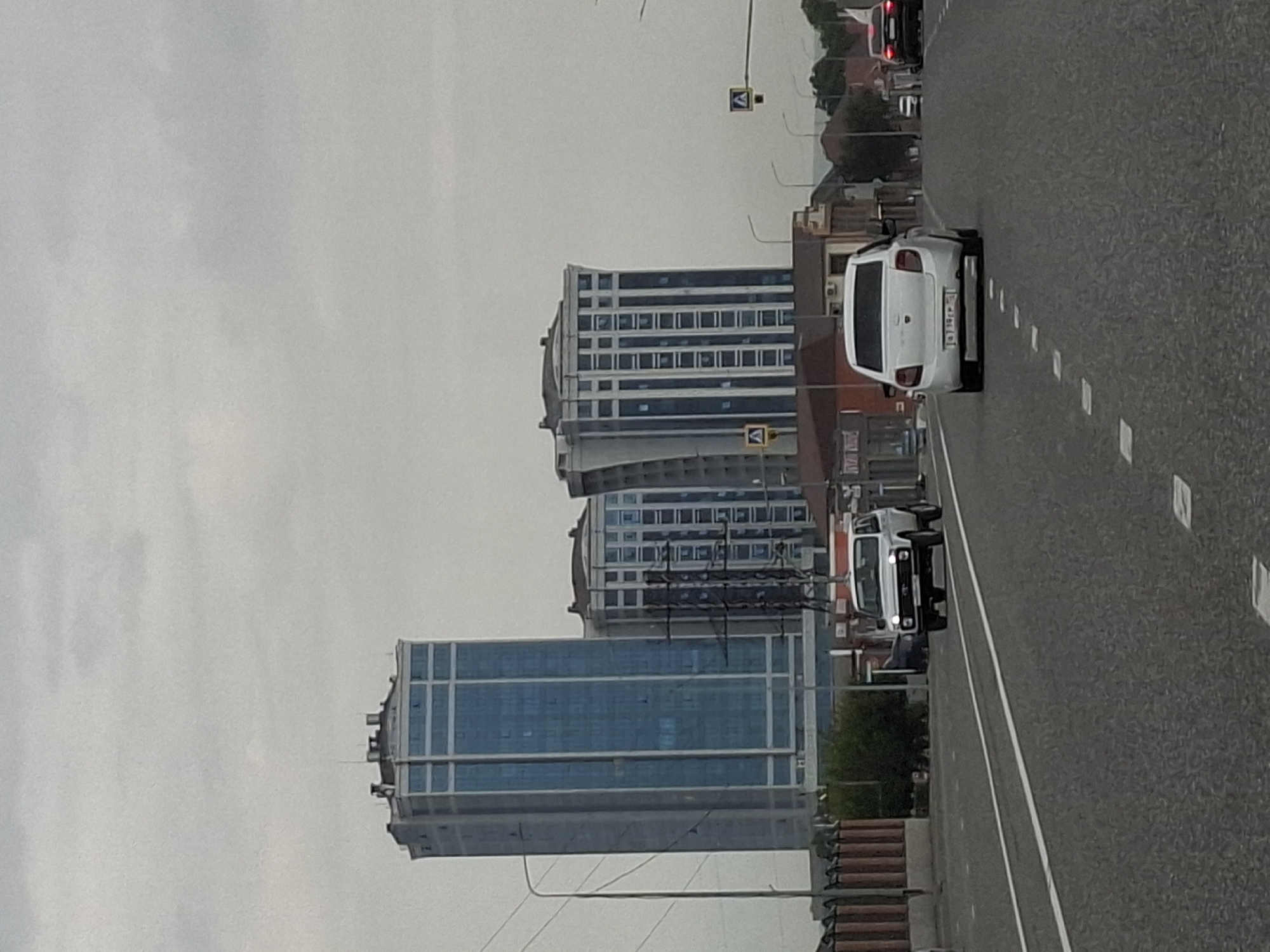 This is a vertical, eye-level, medium shot of a city street in Argun, Russia, on a cloudy day. The scene is dominated by two modern, multi-story buildings with blue glass facades and a gray overcast sky. The buildings are contemporary in design, with repeating patterns of windows. In the foreground, a gray asphalt road with white lane markings stretches into the distance. Several vehicles are visible on the road, including a white car with its license plate visible, and a white van with its headlights on. The overall mood is somewhat subdued due to the cloudy weather. There are no people clearly visible in the image. The time of day appears to be daytime, but the overcast conditions obscure the direct sunlight.