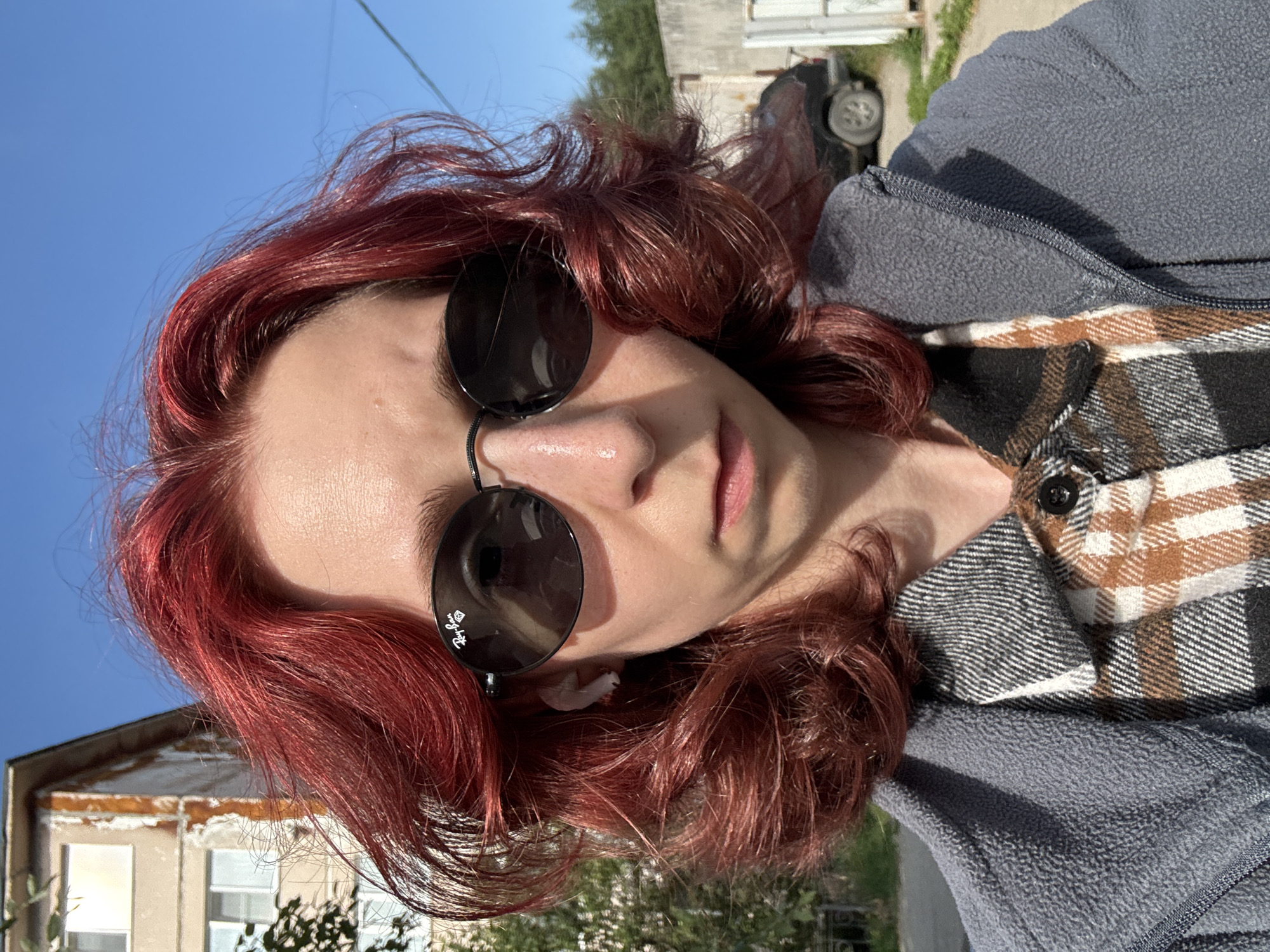 A medium close-up frontal shot shows a person with bright red hair, wearing dark oval sunglasses, a grey fleece jacket, and a plaid shirt underneath, standing outdoors in front of a building. The person is facing the camera, and their head is slightly tilted. The background shows a somewhat dilapidated building with peeling paint, trees, a street, a car, and a concrete wall. The sky is clear and blue. The person is wearing a white earbud in their left ear.