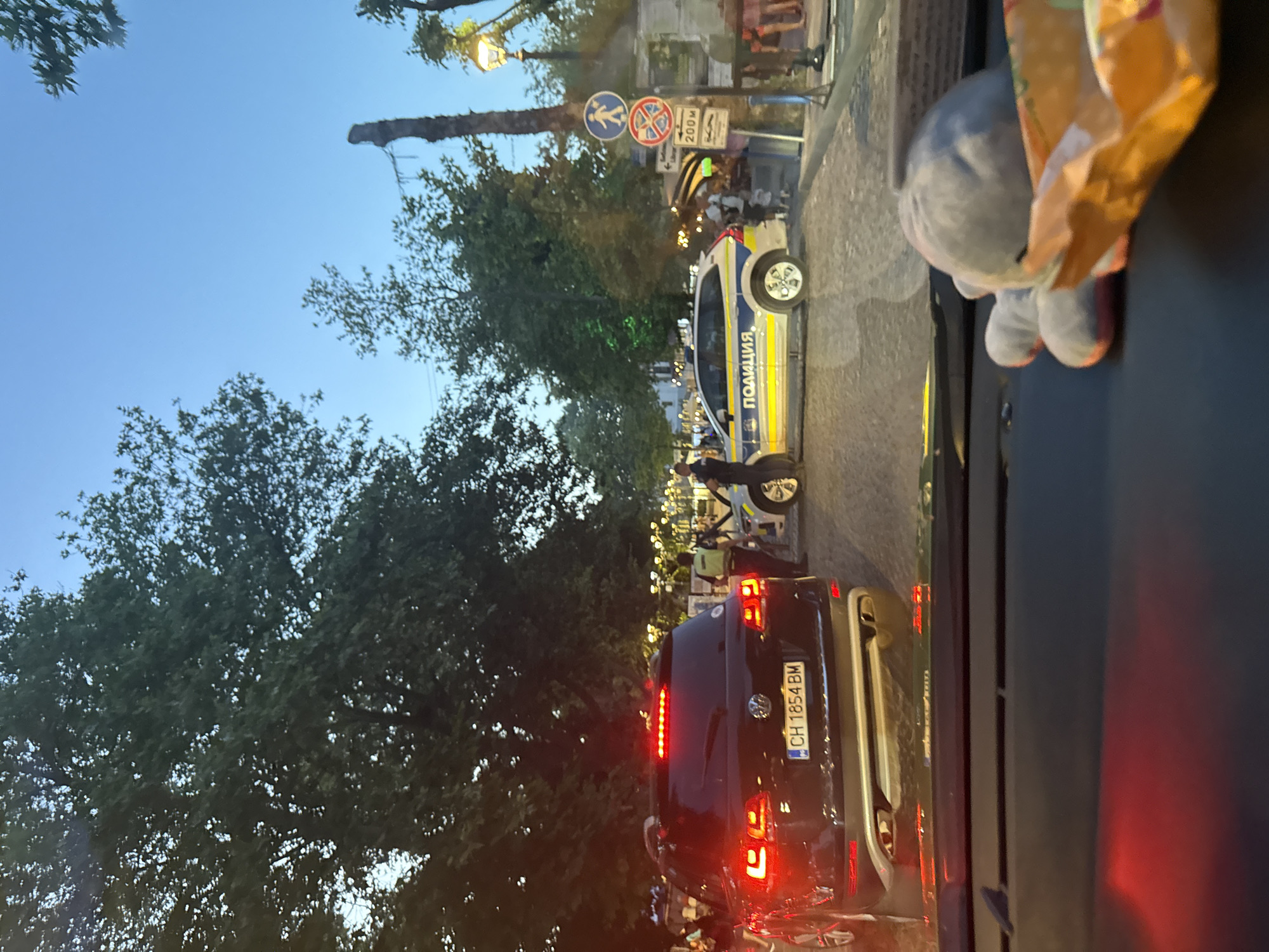 The image depicts a street scene at dusk in Sozopol, Bulgaria. A black Volkswagen SUV with license plate "CH 1854 BM" is stopped, its brake lights illuminated. To the right, a white police car with "ПОЛИЦИЯ" (Police) in blue and yellow lettering is also present. Two individuals in police uniforms are visible near the police car. In the foreground, the interior of a vehicle is visible, with a stuffed toy on the dashboard. Trees line the street, and streetlights are illuminated, casting a warm glow. In the background, there are signs, including one indicating "200M". The overall impression is of a traffic stop or some form of police activity on a paved street.