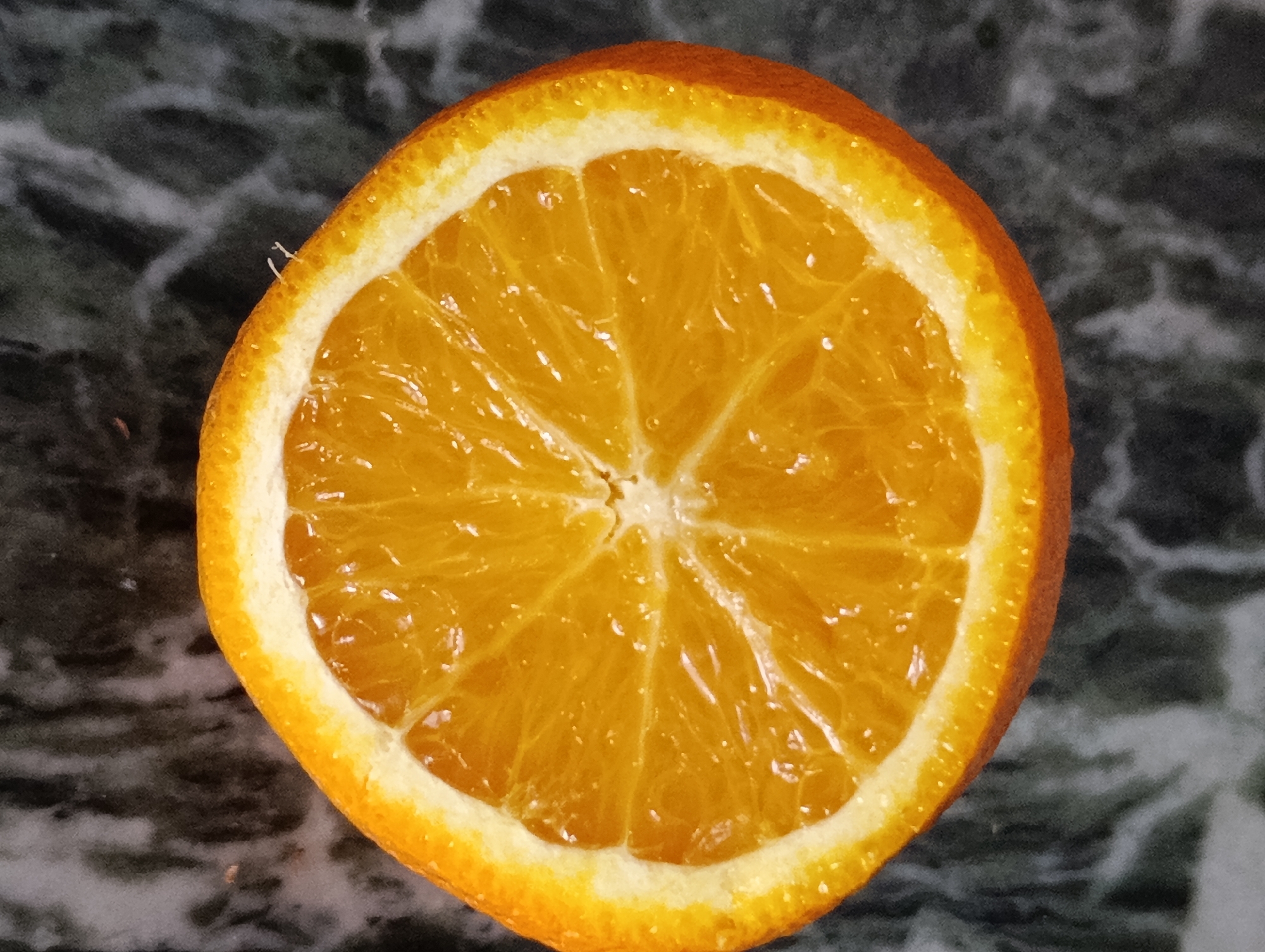 A close-up, overhead shot of a single slice of orange is centered on a dark, mottled surface. The orange slice is vibrant orange, with clearly defined segments and a thin white pith surrounding the pulpy interior. The rind is a textured, darker orange. Tiny, white, hair-like filaments are visible on the outer edge of the orange slice near the top left. The background surface has a marbled appearance with streaks of dark gray and black, resembling granite or a similar stone. The lighting is direct, casting subtle shadows and highlighting the glossy texture of the orange. There is no indication of the city of Vashisht, India, in this image.