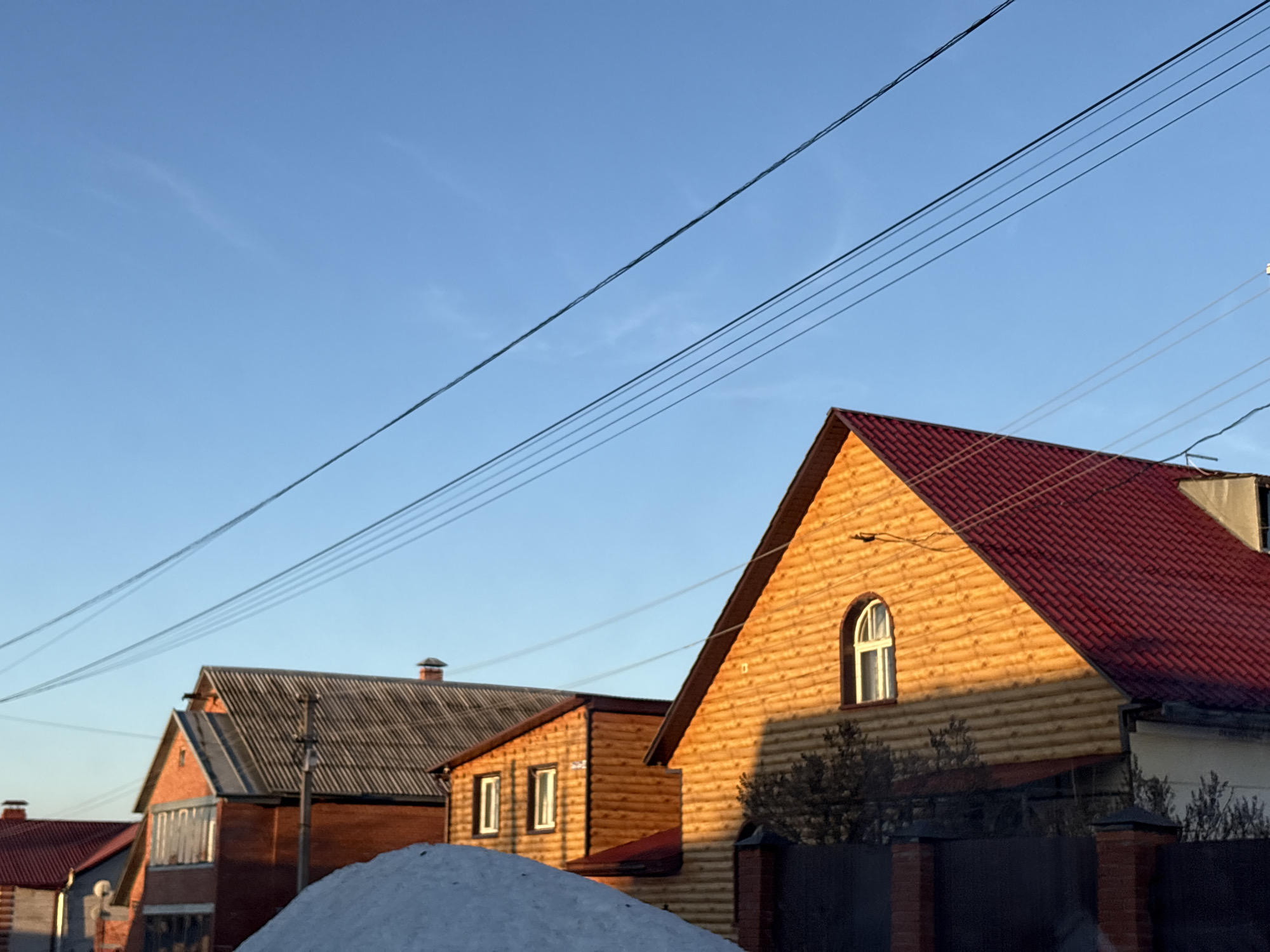 The image depicts a neighborhood in Прикамский, Russia. The scene shows a row of houses under a clear blue sky with several electrical wires running overhead. There are at least three houses visible, each with distinct architectural styles. The house on the right has a light brown wooden exterior with a dark red tiled roof and a single arched window near the peak. The house in the center has a light brown wooden exterior, with windows and a reddish tile roof. The house on the left is constructed from red brick with a gray tiled roof. A pile of snow sits in front of the houses. A dark fence is also visible. The lighting suggests it is early morning or late afternoon due to the long shadows.
