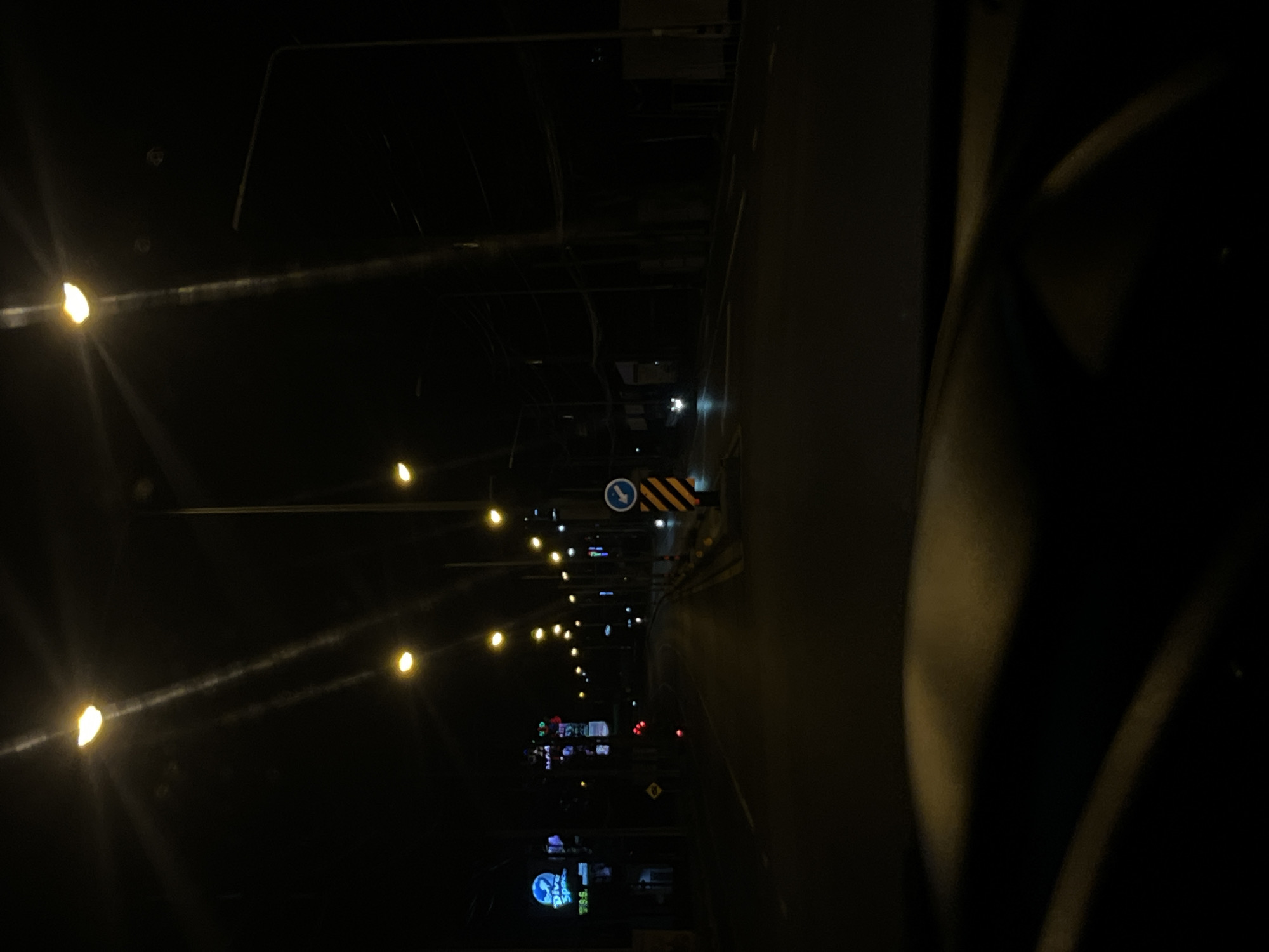 The image depicts a street scene at night in Chalong, Thailand. The street is illuminated by numerous streetlights and neon signs. On the left side of the road, there is a neon sign that reads "Dive Spot." Further down the road, there are more buildings with illuminated signs, some of which appear to be restaurants or shops. The road itself is dark and appears to be wet, possibly from recent rain. There are no people visible in the image, and only a few vehicles are seen in the distance, with their headlights on. The overall atmosphere is dark and somewhat deserted, typical of a quiet street in a Thai city at night. The perspective suggests the photo was taken from inside a vehicle, looking out.