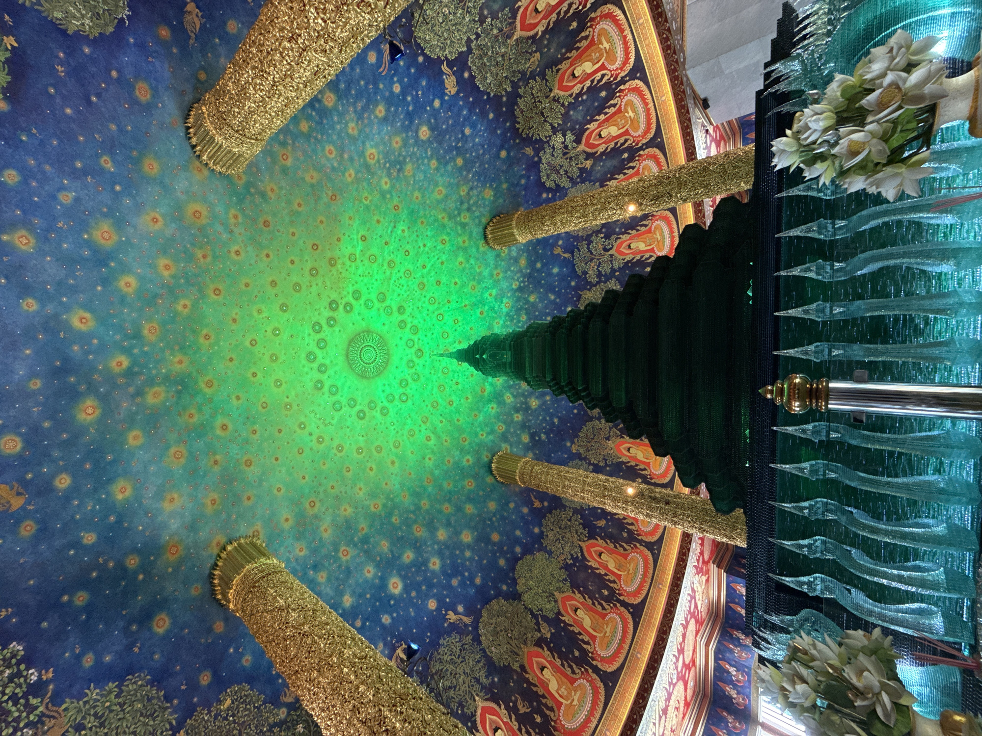 The image captures the ceiling of Wat Arun Ratchawararam Ratchawaramahawihan (Temple of Dawn) in Bangkok, Thailand. The ceiling is painted with a gradient that shifts from a deep blue at the periphery to a vibrant green at the center. Small, circular patterns resembling stars or floral motifs are scattered across the entire surface. Four ornate, golden pillars support the structure, two on each side of the image, and appear to be constructed from intricately carved metal. Painted along the ceiling's edge are depictions of reclining Buddha figures. A partially obscured section on the right includes what appears to be glass ornaments and white artificial flowers.