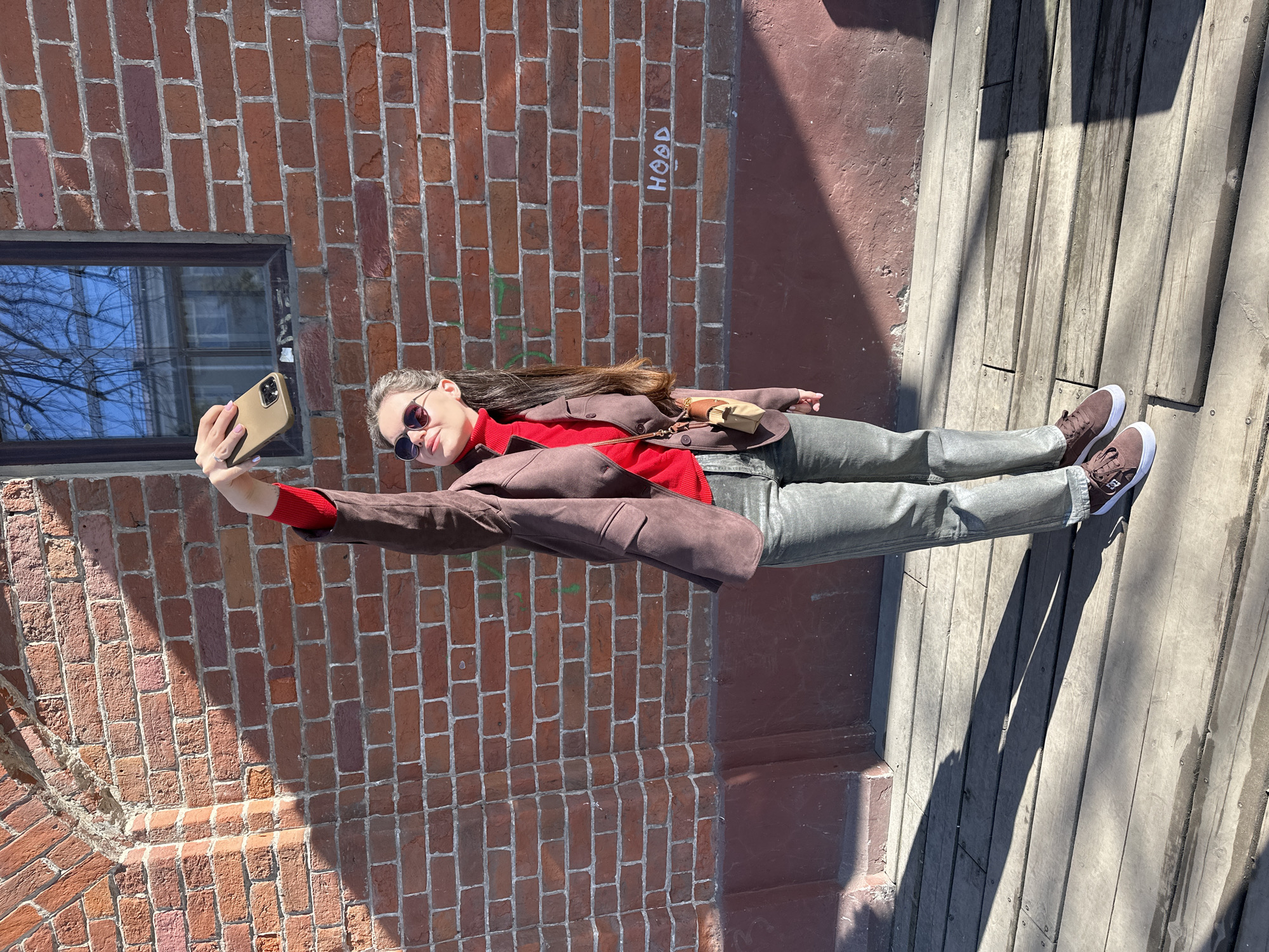 A person is standing outdoors, posing for a selfie against a brick wall and a wooden structure that appears to be stairs. The person is wearing sunglasses, a red turtleneck, a brown jacket, and gray pants. They are holding a smartphone to take the photo. The brick wall has a window and the word "HOOD" written on it. The wooden stairs are weathered. The location is Kazan, Russia.