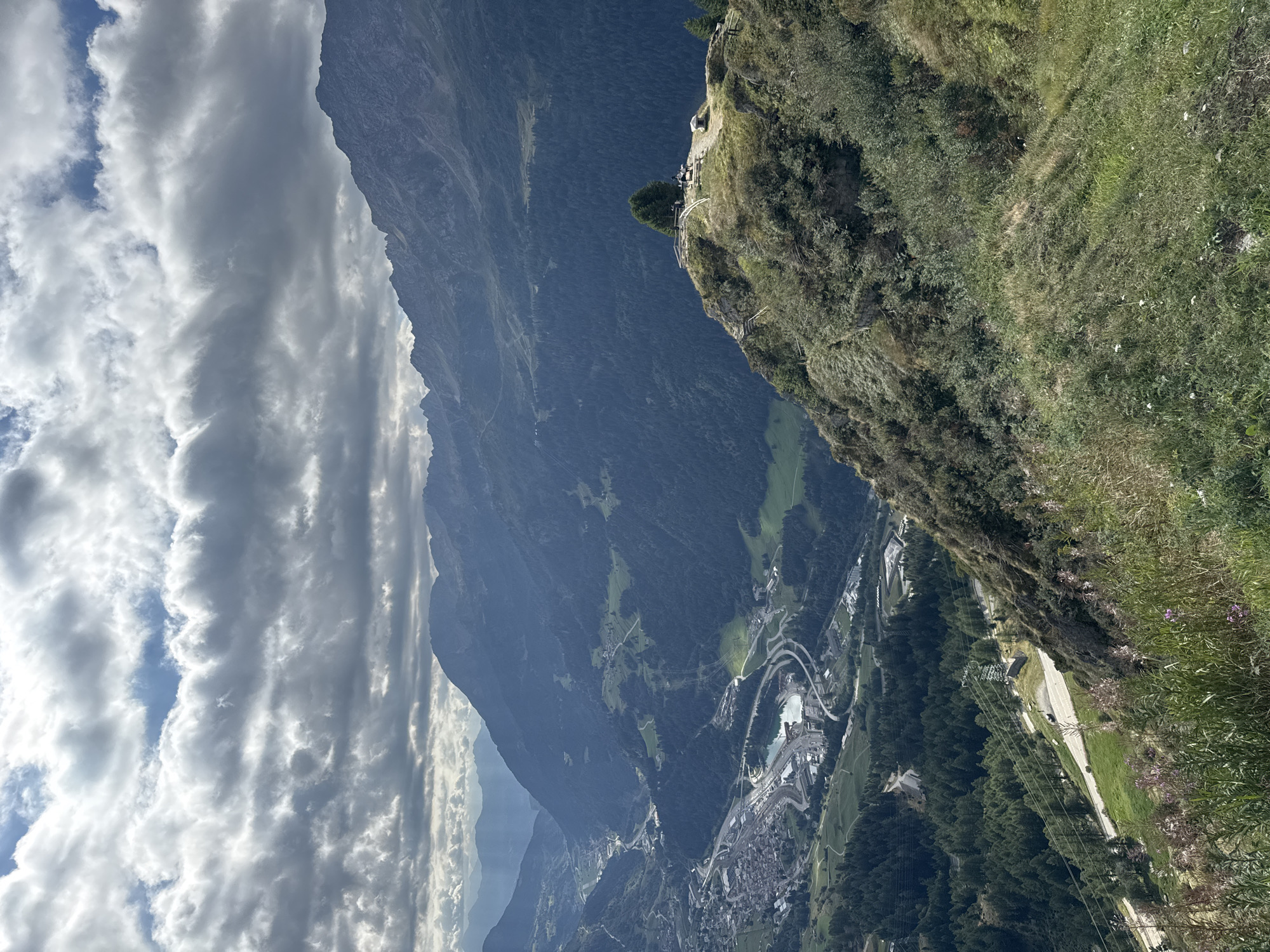 This is a high-angle, medium shot of a mountainous landscape with a valley below. The sky is filled with dramatic, cloudy formations, with patches of blue sky visible. In the foreground, a steep, grassy slope is covered in wildflowers and dry grasses. A winding road cuts through the midground, bordered by a dense forest of evergreen trees.
Further down in the valley, a town is nestled, with buildings and infrastructure visible. A body of water, possibly a lake or reservoir, is surrounded by roads and buildings. A highway interchange is also prominent in the valley. In the background, multiple layers of mountains recede into the distance, their peaks softened by the atmospheric haze.
On a rocky outcrop in the mid-right foreground, a small group of people are gathered around a tree. They appear to be standing or walking, observing the view. A wooden railing is present on the outcrop, indicating it is a designated viewpoint. The overall impression is one of a vast, natural landscape with human settlement integrated into the valley. The location is Airolo, Switzerland, likely captured from a high vantage point overlooking the town and surrounding mountains.