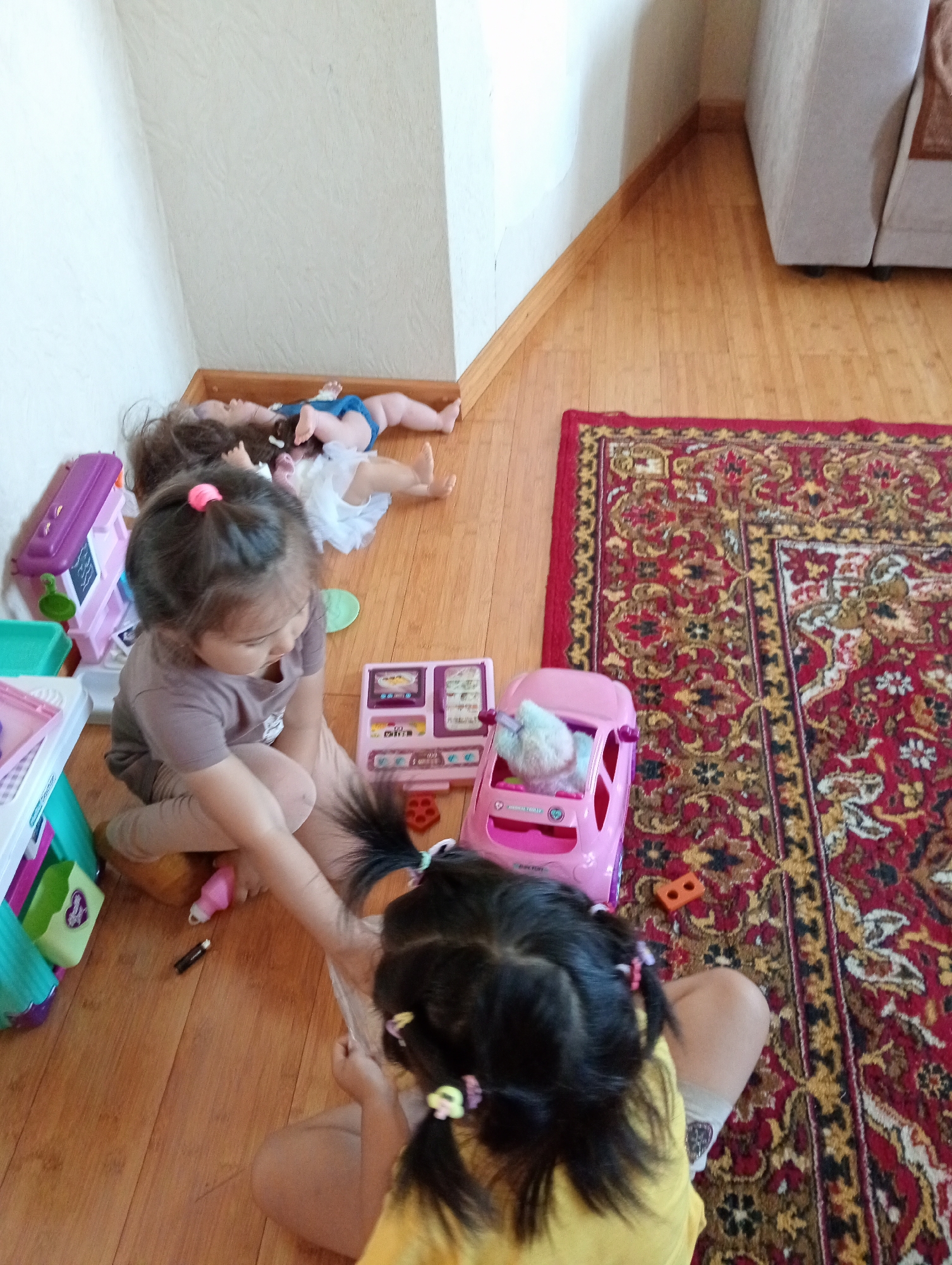 The image depicts two young girls playing on a wooden floor. In the background, two baby dolls are lying on the floor near a wall. In the foreground, one girl is sitting and playing with toys, while the other girl is also on the floor, seemingly engaged in play. Various toys, including a pink toy car and a toy kitchen, are scattered around the children. The room appears to be a living space, with a large red patterned rug on the floor. A couch is partially visible in the upper right corner. The scene is illuminated by natural light, casting shadows on the floor. The environment is consistent with an indoor residential setting in an unspecified location within Kazakhstan.