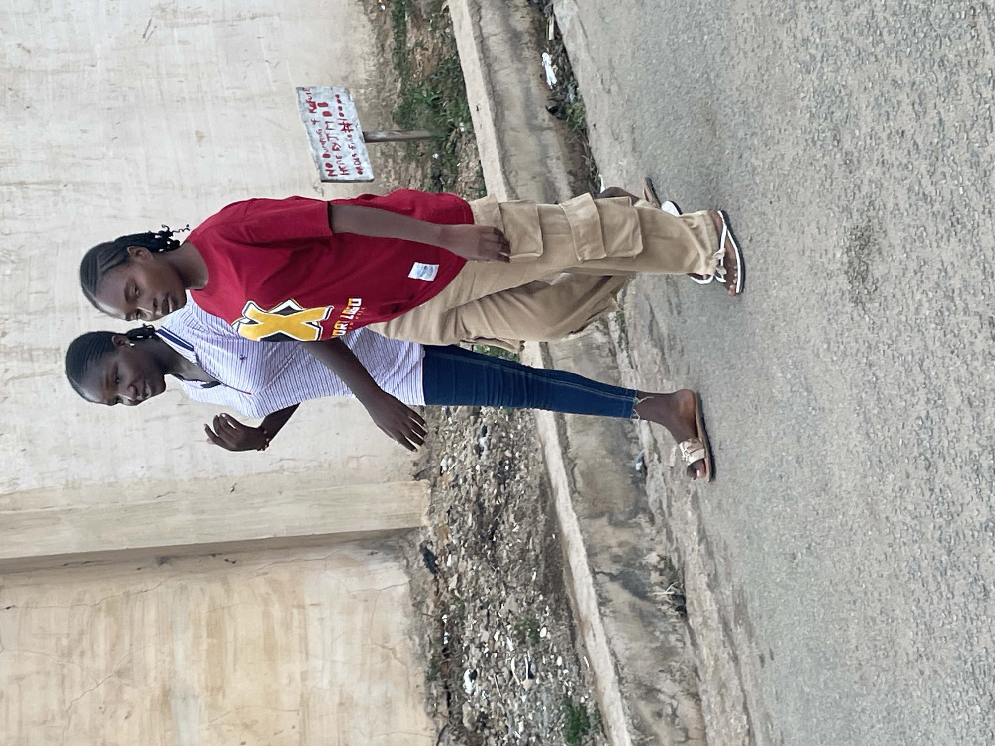 Two young women are depicted walking or standing casually on a paved surface alongside a light-colored concrete wall in what appears to be an urban or semi-urban setting in Jos, Nigeria. The image is originally oriented vertically, but when viewed upright, the subjects move from left to right.
The woman in the foreground, positioned on the left, wears a light blue and white striped long-sleeved button-up shirt, dark blue jeans, and light-colored sandals. Her left arm is slightly bent, and her hand is open. Her hair is styled in dark braids.
Slightly behind and to her right is the second woman, dressed in a red short-sleeved t-shirt featuring a prominent yellow 'X' logo and partial text, possibly "COLLEGE," on the front. She wears loose-fitting khaki cargo pants and brown and white sandals. Her hair is also braided.
The environment includes a distressed, light-colored concrete or stucco wall on the left, exhibiting cracks and signs of wear. The ground is a light gray paved surface, likely asphalt or concrete. To the right of the women, between the paved area and a raised curb or another concrete structure, lies a strip of uneven ground covered with dirt, small rocks, and sparse debris.
A small, rectangular white sign, attached to a pole, is visible behind the women and closer to the right wall. Red handwritten text on the sign is discernible, reading: "NO BURNING & RUBBISH" and "N10,000 FINE." This indicates a prohibition against burning refuse, enforceable with a penalty of ten thousand Nigerian Naira.
The scene occurs during the daytime under bright, even lighting, suggesting either clear skies or an overcast condition due to the absence of harsh shadows. The women's facial expressions appear neutral.