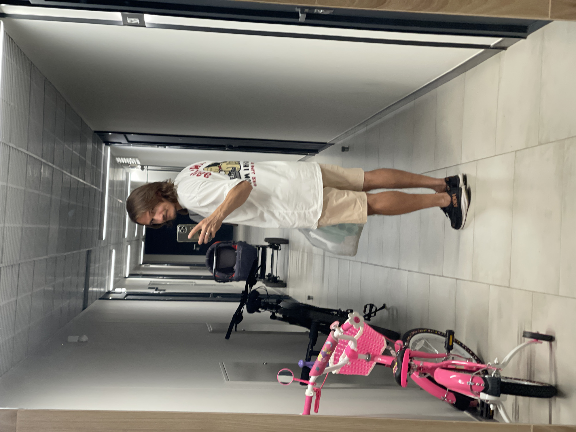 A man is taking a mirror selfie in a well-lit, modern apartment building hallway. He is wearing a white t-shirt with a graphic print on the back and light-colored shorts, along with black and white athletic shoes. In the foreground to his left, a pink child's bicycle with training wheels is parked. Behind the bicycle, a dark-colored stroller is visible. The hallway has a tiled floor and white walls, with fluorescent lighting panels on the ceiling. The man is holding a smartphone in his right hand, capturing the reflection.