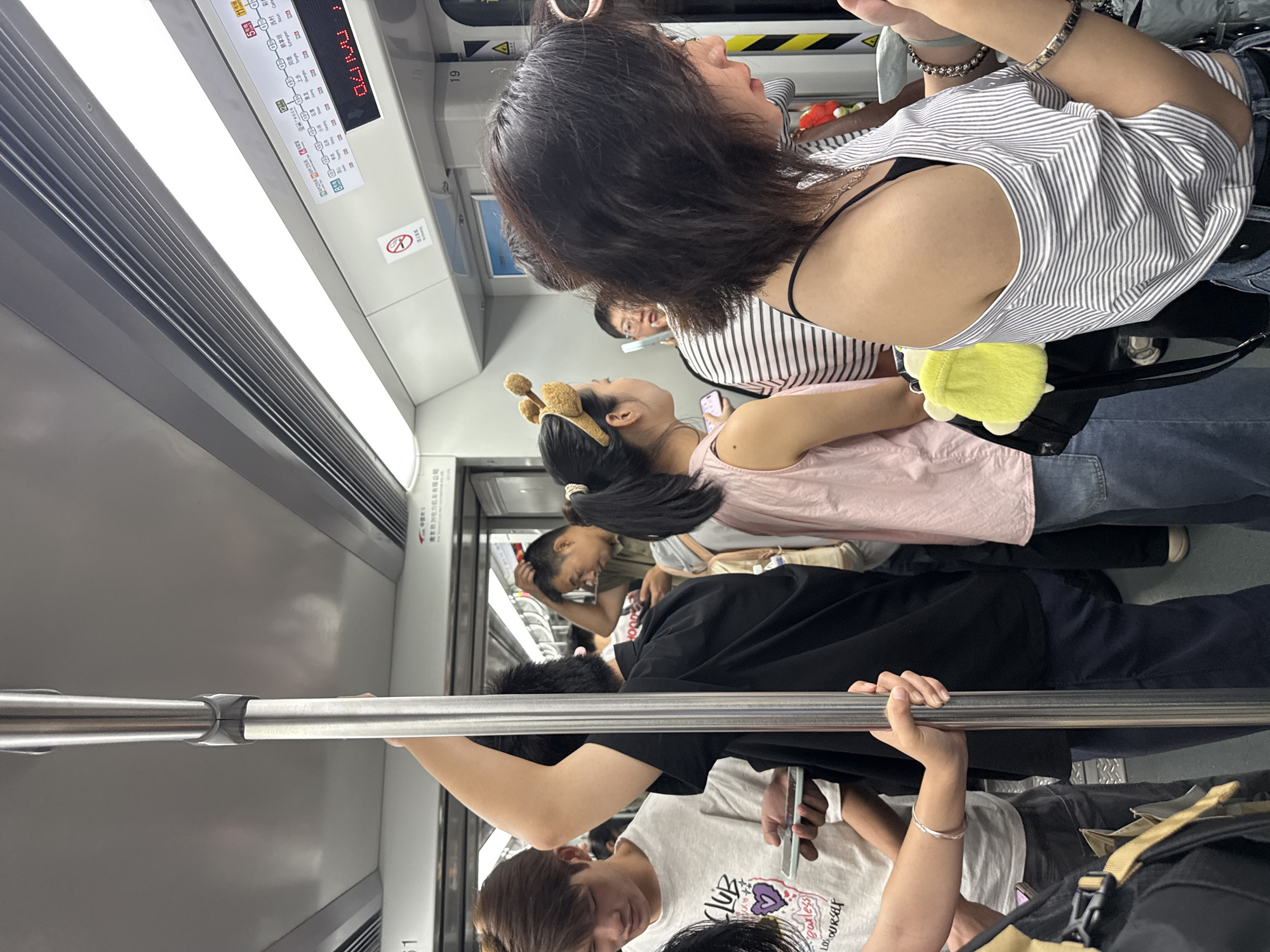 The image depicts the interior of a crowded subway train, likely in Haizhu District, China, based on the visible signage. Several individuals are standing, holding onto vertical poles and handrails for support. The lighting is artificial, originating from fluorescent tubes overhead. In the foreground, a woman with short, dark hair wearing a horizontally striped off-the-shoulder shirt is visible from the chest up. To her left, a woman with dark hair tied back and wearing a light pink sleeveless top and denim jeans is also visible, with a fuzzy yellow accessory attached to her backpack. Further to the left, a young man in a grey t-shirt with graphics is looking down at his phone. Other passengers are partially visible in the background, with one man in a black t-shirt holding a vertical pole and another man with short hair and a light brown shirt adjusting his hair. A digital display above the windows shows route information in Chinese characters. The overall impression is one of a busy public transportation environment.