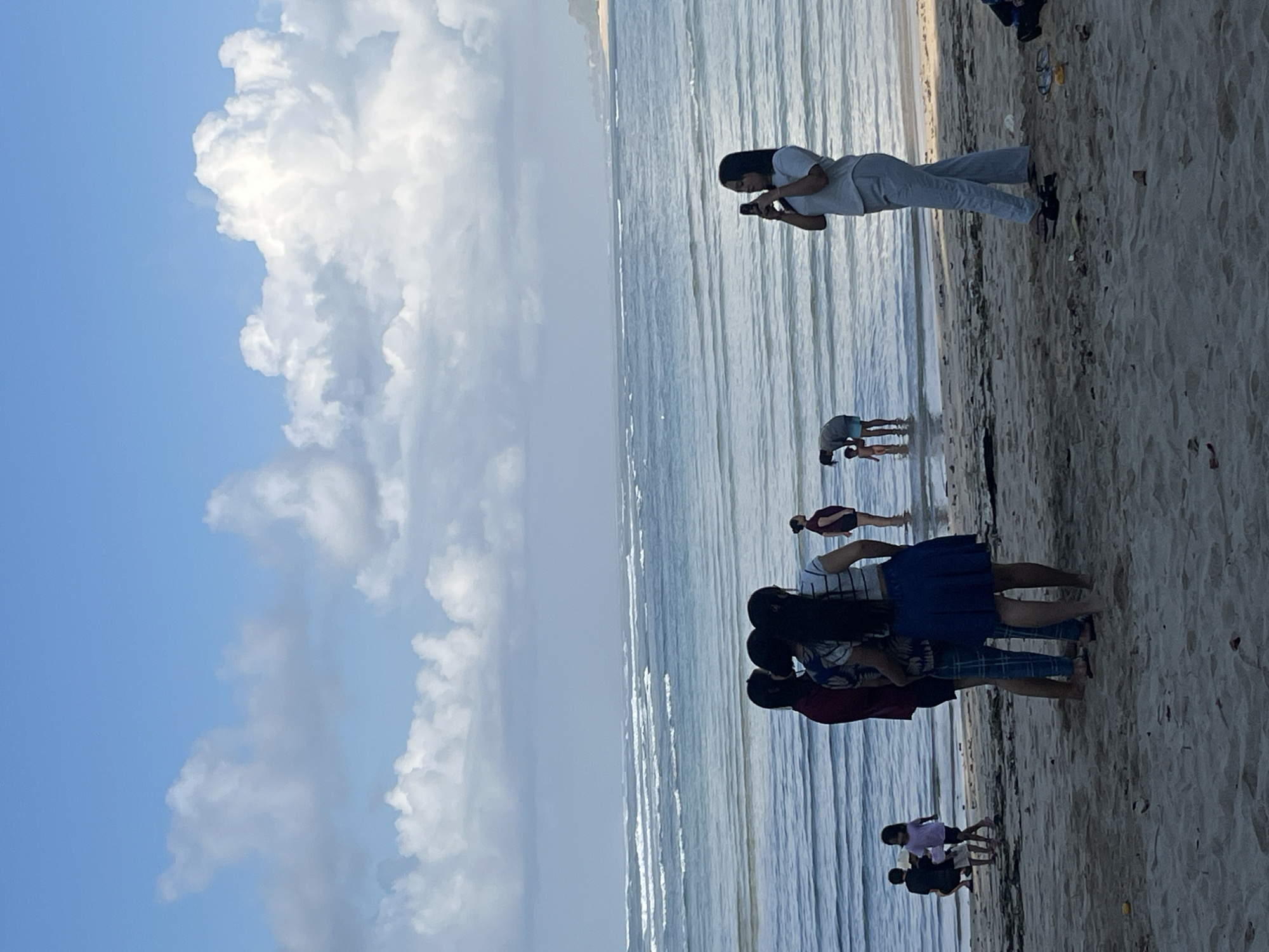 In Kuta Selatan, Indonesia, a group of people are gathered on a sandy beach with the ocean in the background. The sky is a clear blue with scattered white clouds. The ocean is calm with small waves rolling in. In the foreground, on the sand, several individuals are standing and interacting. Three women are grouped together, facing away from the camera, with one wearing a red shirt, another a patterned top, and the third a striped shirt and a blue skirt. To their left, a child in a pink shirt and shorts is standing, and a person in a dark shirt and shorts is crouching. Further to the right, a woman in a light gray top and pants is holding up a phone, appearing to take a photo. In the shallow water near the shore, a person in a dark top and shorts is bending over, and a young child in a bathing suit is standing nearby. In the distance, the coastline curves, with vegetation visible on a hill. The beach itself is sandy, with some scattered debris and foliage.