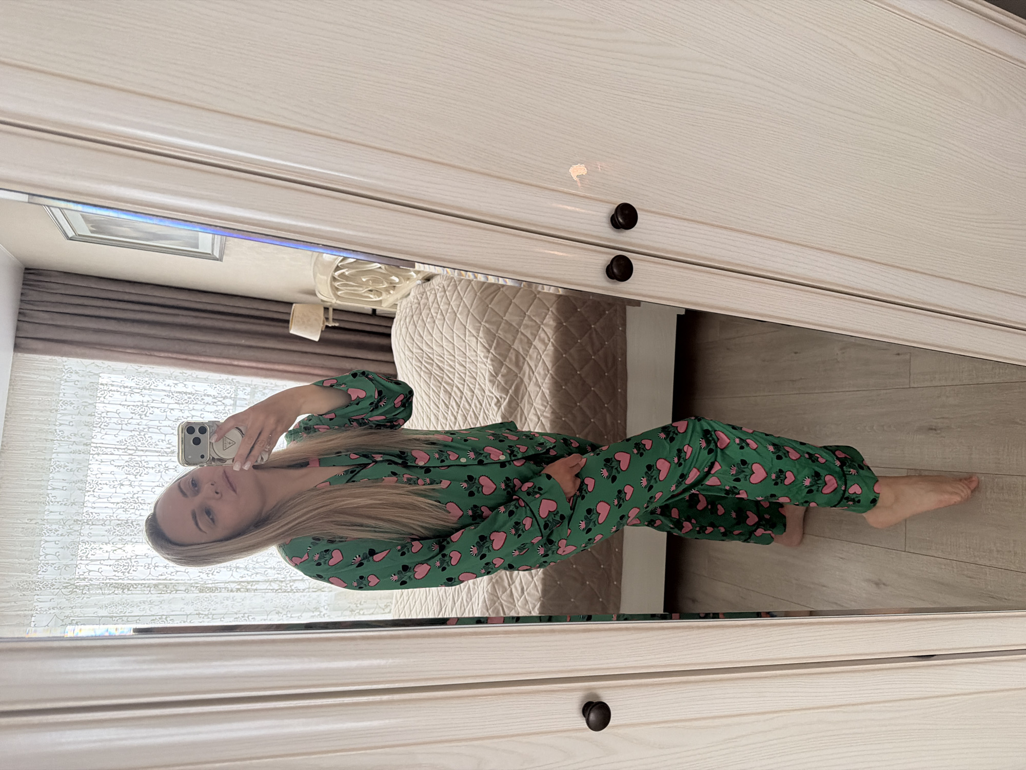 A person takes a mirror selfie in what appears to be a bedroom in Votkinsk, Russia. The individual, with long, straight blonde hair, is dressed in a two-piece set of green pajamas adorned with a playful pattern of pink hearts and black paw prints. They are holding a smartphone, with a distinct camera array and a grip accessory, to capture their reflection. The person is barefoot.
The mirror, which occupies the majority of the frame, is set into a light-colored wooden wardrobe or cabinet. This piece of furniture features paneled doors and dark, round knobs. The reflection reveals a well-appointed bedroom environment.
In the background, a bed with a light-colored, quilted headboard and matching bedding is visible. To the left, a window is partially obscured by decorative white lace curtains and plain brown drapes, suggesting daytime. A framed picture hangs on the wall above the bed, and a lamp with a white shade rests on a bedside table. The room has a light wooden floor. The person's expression is neutral. No discernible text is present on any objects or clothing.