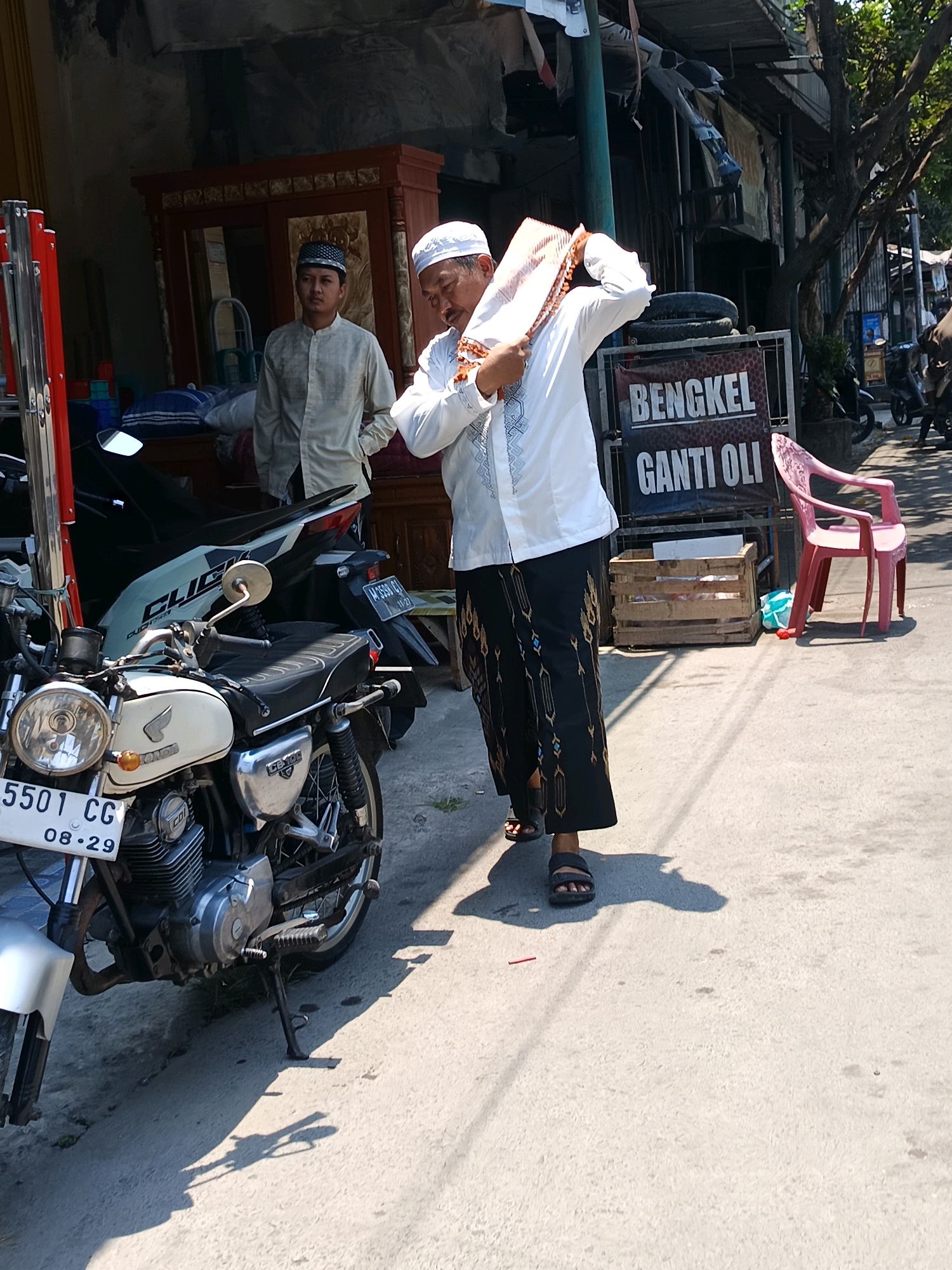 In Blega, Indonesia, an older man wearing a white shirt and a peci cap is adjusting a patterned cloth over his head. He is standing next to a parked motorcycle. Behind him, another man in a light-colored shirt and peci cap stands near a wooden cabinet. The scene appears to be outdoors, with a building in the background and a pink plastic chair visible. A sign that reads "BENGKEL GANTI OLI" is also present. The motorcycle in the foreground has a license plate that reads "5501 CG 08-29". The lighting suggests it is daytime.