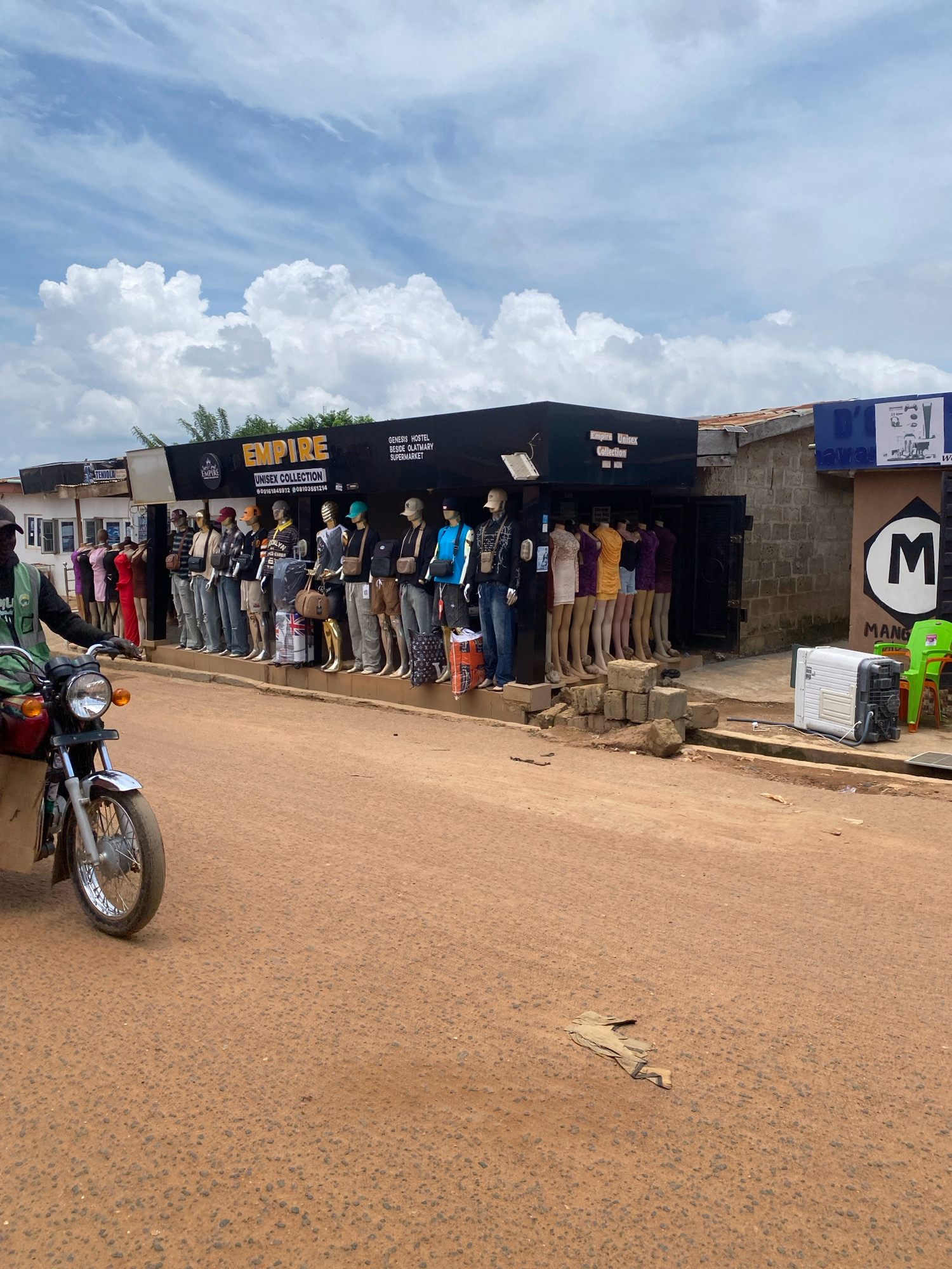 In Iworoko, Nigeria, a street scene unfolds under a cloudy sky. A dirt road, marked by tire tracks and scattered debris, leads past a row of shops. One shop, prominently displaying "EMPIRE UNISEX COLLECTION," features a line of mannequins dressed in various apparel, from casual shirts and jeans to more formal outfits. Another shop to the right has a sign with the letter "M" in a circle and the word "MANGO" below it. A motorcycle, ridden by a person wearing a green jacket and a cap, is positioned on the left side of the road, partially obscuring the view of the shops. In front of the "EMPIRE" shop, a white portable air conditioner unit rests on the ground next to stacked bricks. The environment appears to be a busy commercial area with a mix of building materials and a sense of everyday life.