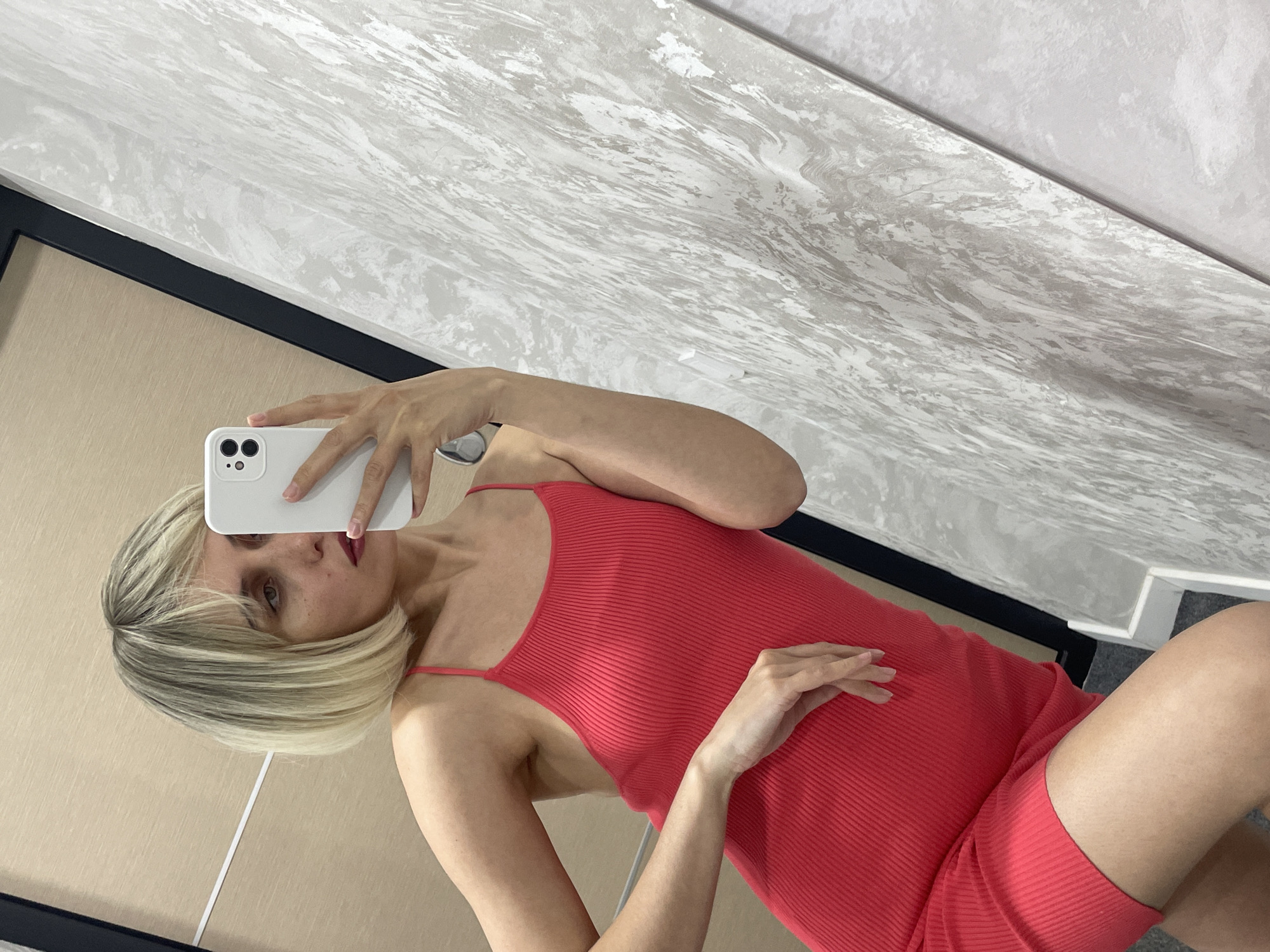 The image is a mirror selfie featuring a woman with short, blonde, bob-cut hair. She is wearing a form-fitting, ribbed, red spaghetti-strap dress or top with matching shorts. Her left hand holds a white iPhone with a triple-camera setup, angled to capture her reflection. Her right hand is gently placed on her stomach or hip area. She wears red lipstick and looks towards the phone's screen.
The background reflected in the mirror consists of a light wood-toned panel or wall on the left side, featuring a thin vertical white strip. A thin black frame or trim separates this wood-toned section from a light grey textured surface that occupies the upper and right portions of the reflection. This grey surface exhibits a swirling or marbled pattern. The entire image is oriented diagonally, suggesting an angled pose or a rotated photograph.
There are no specific cues for the time of day or weather, as the scene appears to be indoors. The woman's expression is neutral, and no visible text is present in the image. Based on the provided context, the location is Izhevsk, Russia.