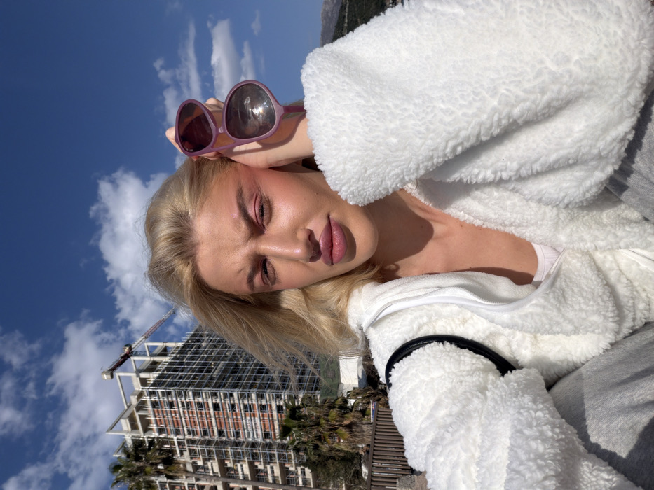 An outdoor selfie captures a woman with blonde hair and fair skin, illuminated by natural light under a bright blue sky with scattered white clouds. She wears a white fleece jacket over a white top, with grey sweatpants visible below. Her eyes are slightly squinted, and her lips are pursed, accentuated by pink eyeshadow and light pink lipstick. Her raised left hand holds a pair of purple-framed sunglasses against her temple.
The background identifies the location as Budva, Montenegro. To the left, a multi-story building under construction is prominent, featuring visible scaffolding and topped by a tower crane. Several palm trees are positioned in front of and around the construction site. A wooden fence or barrier runs horizontally behind the woman on the left side of the frame. Distant mountains are visible along the far right horizon.