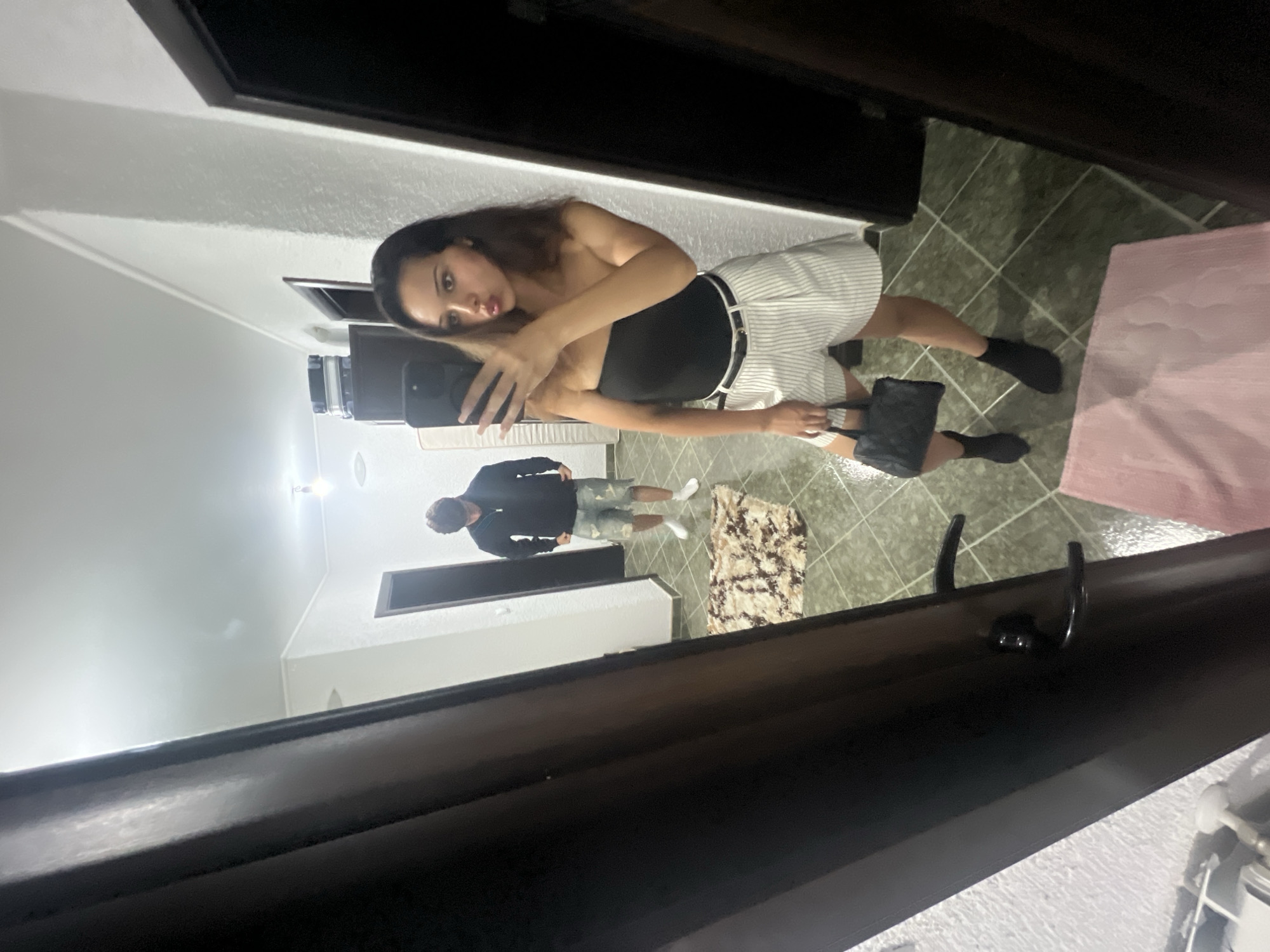 A young woman in a strapless black top and white pinstripe shorts is taking a selfie in a mirror. She is holding a black quilted handbag in her left hand and a phone in her right hand. She is wearing black sock boots. In the reflection of the mirror, a young man in a black zip-up jacket and denim shorts is standing behind her, looking down at his feet. The setting appears to be an indoor hallway or room, with tiled flooring and white walls. There is a rug on the floor and a door visible in the background. A mattress is leaning against the wall to the left. The overall impression is a casual, everyday scene. The image was taken in Zlatograd, Bulgaria.