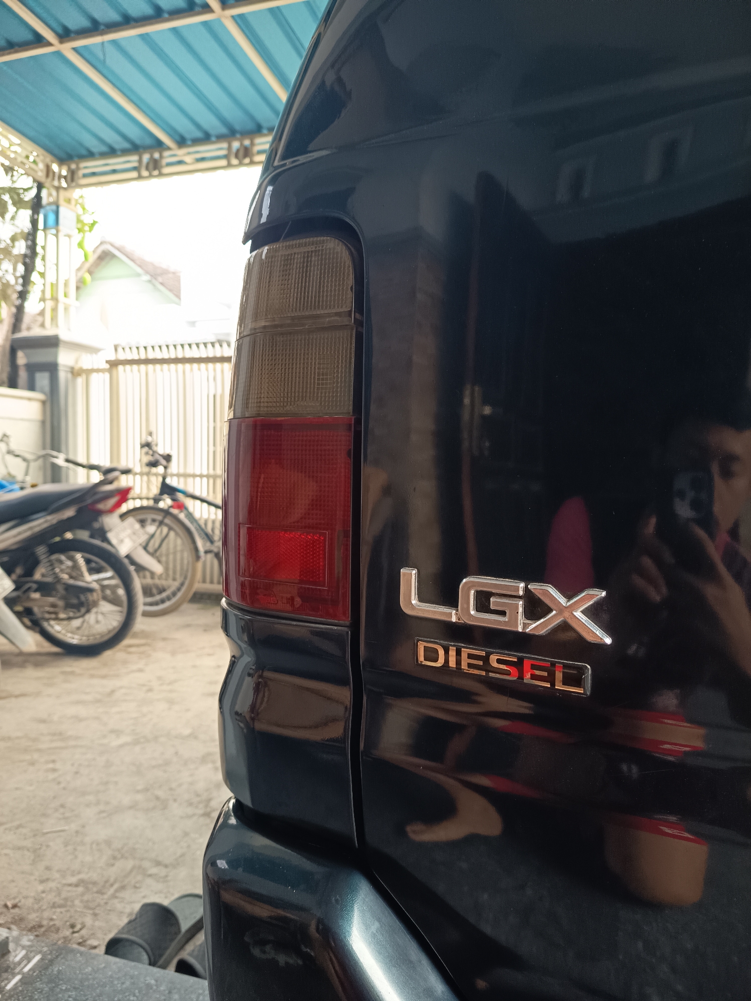 The image captures a close-up, rear-side view of a black vehicle, possibly a van or SUV, parked outdoors under a blue corrugated roof structure. The vehicle's tail light assembly, a red and clear plastic unit, is prominently featured on the left side of the frame. On the right side of the vehicle's rear, metallic badges read "LGX" and "DIESEL" in silver and red respectively. The background reveals a paved area with sand or gravel, a white metal fence, a parked motorcycle and bicycle, and a portion of a residential building. A person's reflection is visible in the shiny surface of the vehicle, holding what appears to be a smartphone. The overall setting suggests a residential or common outdoor area in Sugio, Indonesia.