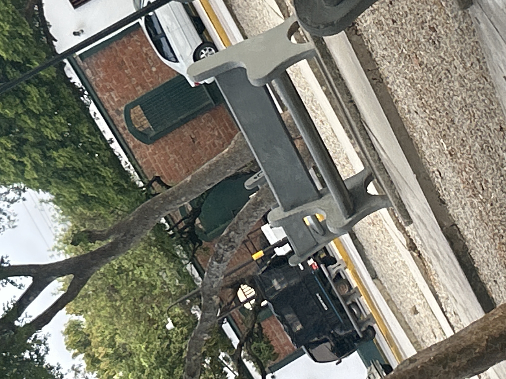 The image depicts a street scene in Santa Ana, Venezuela, with a focus on a grey park bench in the foreground. Behind the bench, a black truck is parked on a street with a yellow line. The truck appears to be a utility or service vehicle. Further in the background, a white sedan is visible parked on the same street. The buildings in the background are constructed of red brick with green architectural elements, possibly doors or shutters. Lush green trees with dense foliage are scattered throughout the scene, indicating a tropical or subtropical climate. The ground is covered with small pebbles or gravel. The sky is overcast. The image is taken from a slightly elevated angle, looking down at the street.