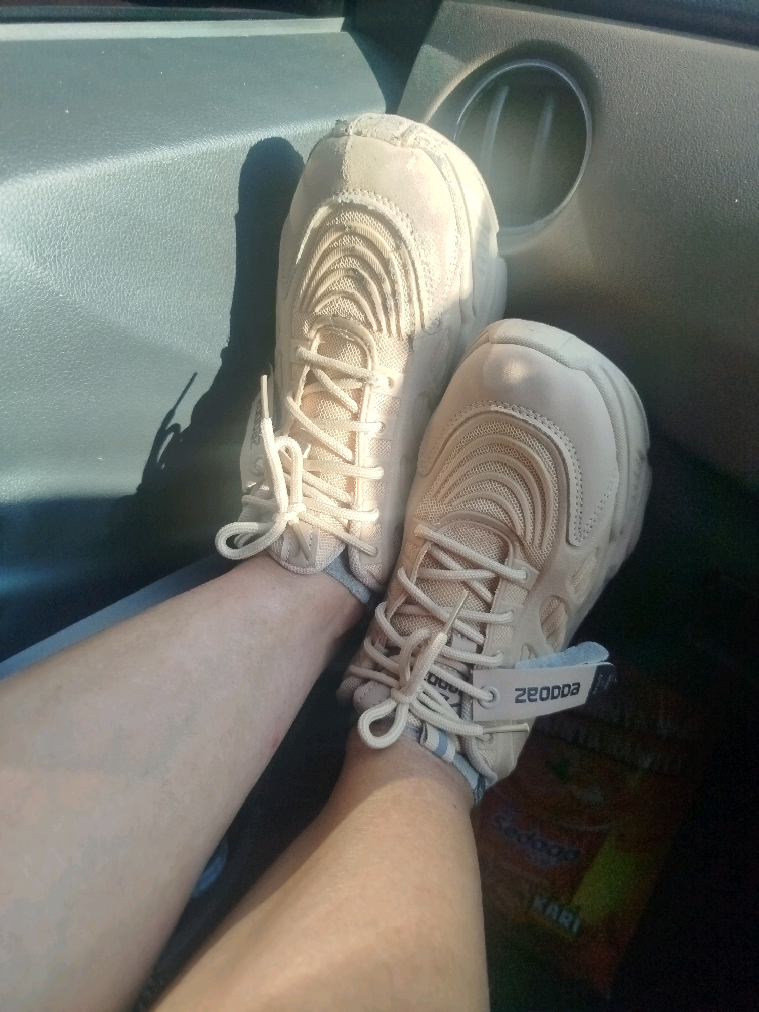 A pair of beige sneakers with white laces is worn by a person whose legs are visible. The sneakers feature a mesh-like design on the toe box and a thick sole. A white tag with the word "ZEODDA" is attached to one of the shoes. The person is seated in what appears to be the interior of a vehicle, with a textured gray dashboard and a circular vent visible. The lighting suggests it is daytime, with sunlight casting shadows. The image is focused on the lower legs and feet of the individual. The location of Suci, Indonesia, is not depicted or discernible within the provided image.