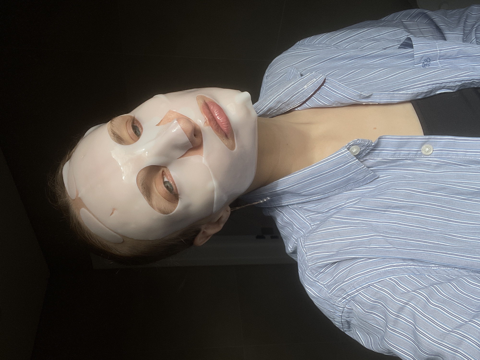 A woman wearing a white sheet mask is reclining. The mask has openings for the eyes and mouth. She's wearing a blue striped button-down shirt over a black top. The location is not identifiable as Подшивалово, Russia from the image provided.
