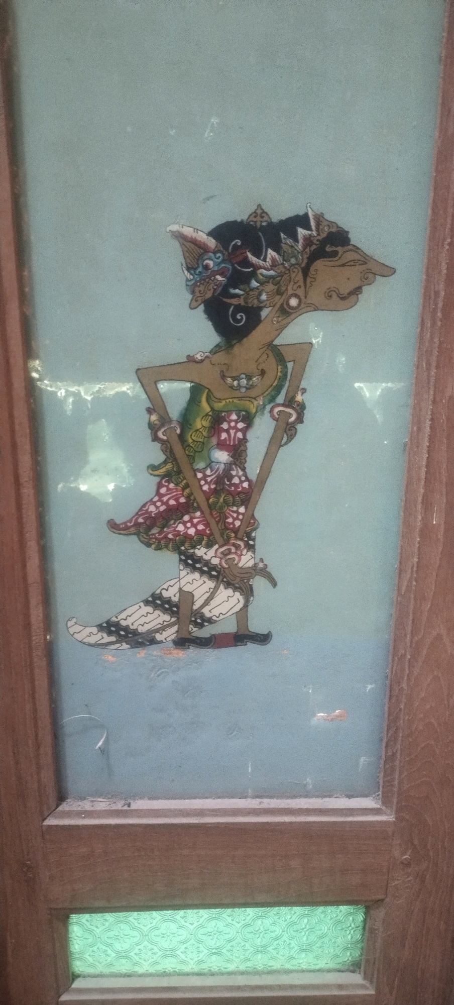 The image shows a traditional Javanese Wayang Golek puppet, a wooden doll with articulated limbs and colorful attire, painted on a glass panel. The puppet is depicted in profile, with an elaborate headdress, facial features, and a decorative costume. The background is a pale blue, and the glass is framed by dark wood. Below the painted panel, there is a section of green patterned glass. The image is taken from a slightly upward angle, with the wooden frame of the window or door occupying the edges of the frame. This object is likely located within a dwelling in Daleman, Indonesia, given the cultural context of Wayang Golek.