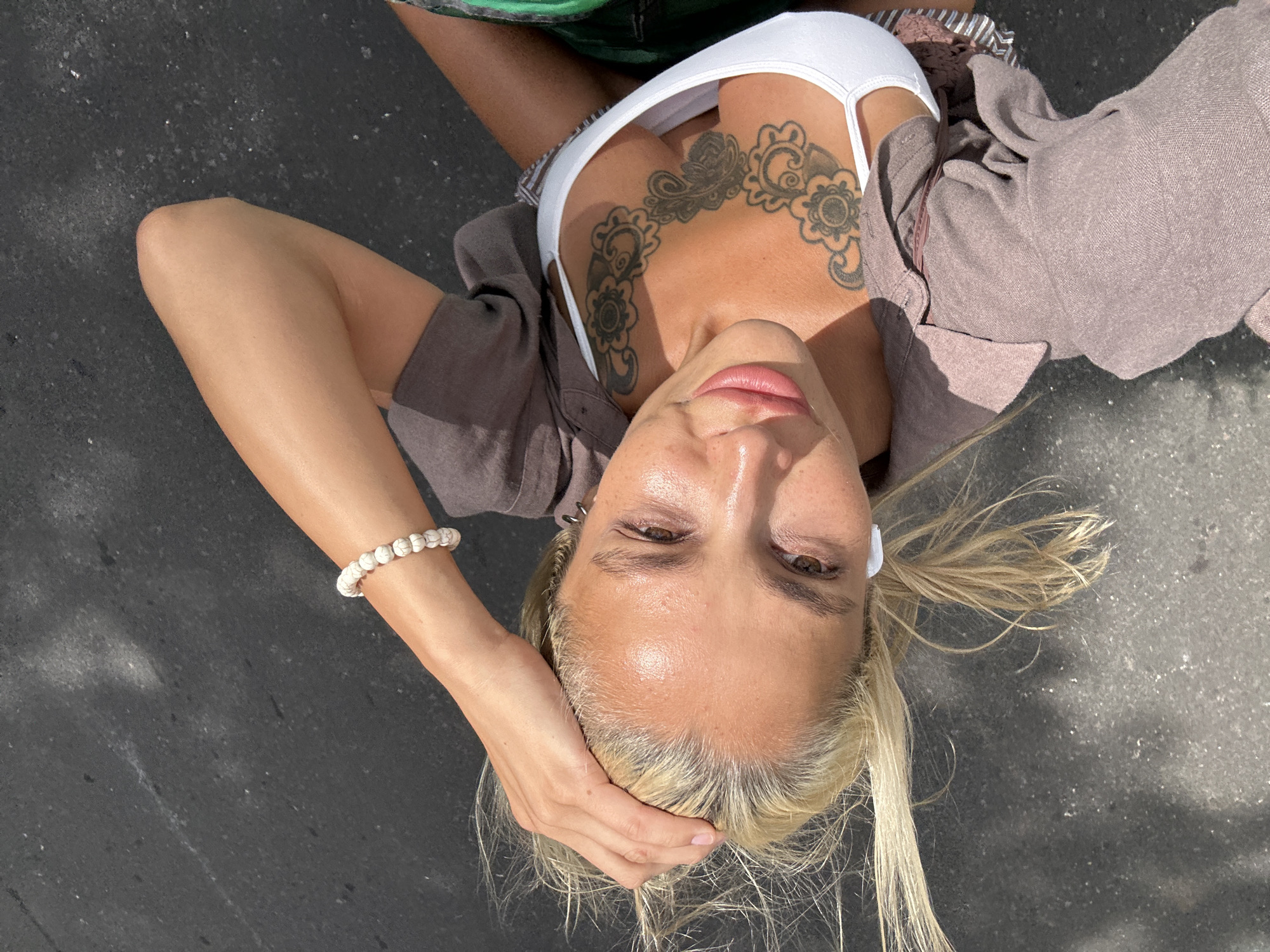 A high-angle, medium shot captures a blonde woman in Kuta Selatan, Indonesia, positioned on asphalt under daylight. She is wearing a white bikini top and a loose, taupe-colored button-up shirt, unbuttoned and draped over her shoulders. Her right hand rests on her head, and she has a tattoo across her chest and upper torso. A white bracelet adorns her left wrist, and she has white wireless earbuds in her ears. Her hair is windblown, and her gaze is directed towards the camera. The background consists of dark asphalt, with scattered light reflections and some fine particulate matter.