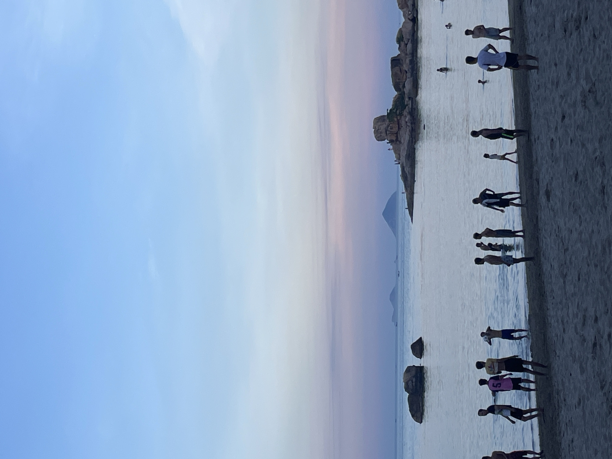The image depicts a shoreline with people, likely taken at Bac Nha Trang Ward, Vietnam. The sky is blue with a gradient transitioning to lighter hues and hints of pink/purple near the horizon. A rocky landmass is visible further out in the water. Several individuals are standing on the beach and in the shallow water. The sand is visible along the right edge.