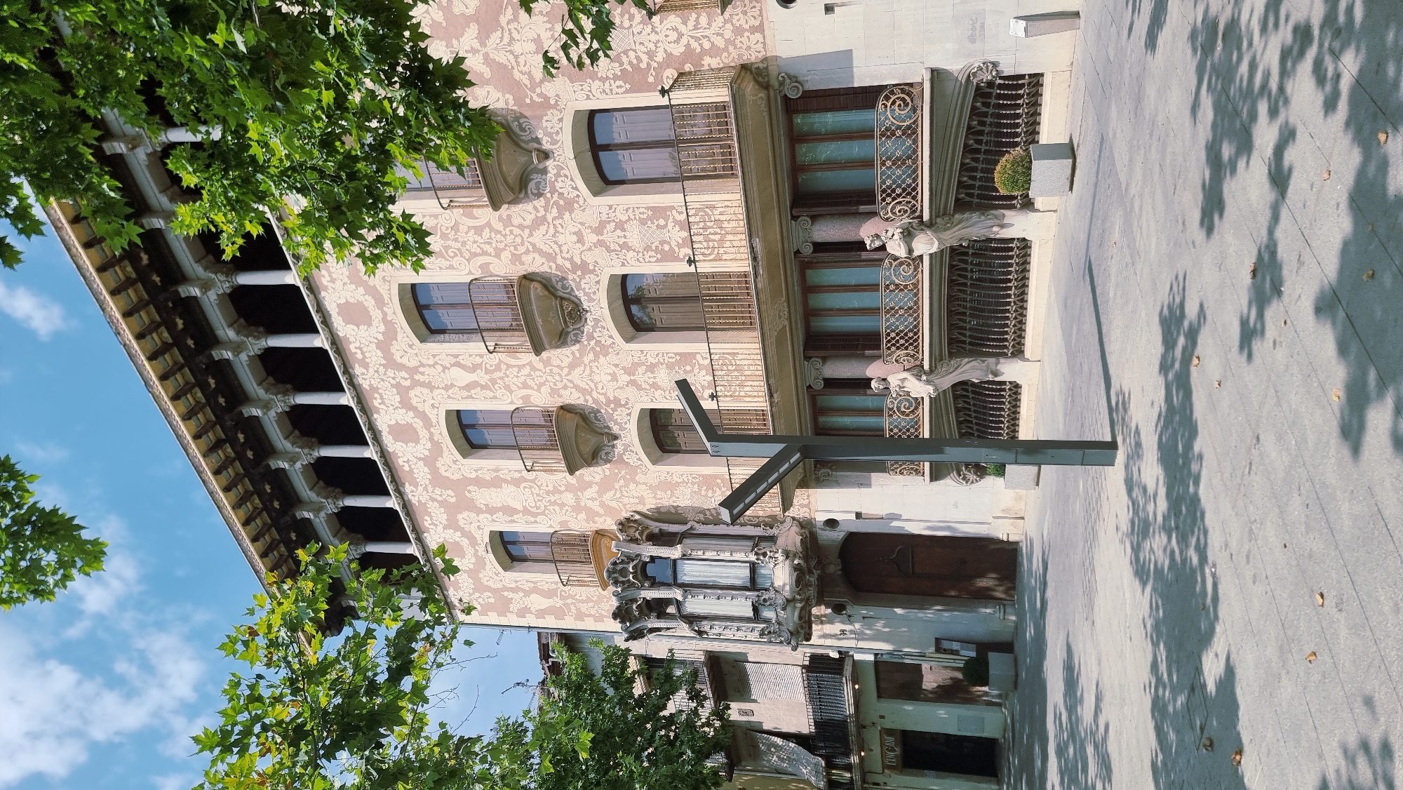The image shows the facade of a building with ornate architectural details, possibly a historic building, in Olot, Spain. The building features elaborate decorative patterns on its exterior, arched windows with decorative balconies and wrought-iron grates, and ornate sculptures adorning the facade. A covered walkway with columns is visible on the left side of the building. Trees with green leaves are in the foreground, casting shadows on the pavement. The sky is blue with scattered clouds, suggesting it is daytime. The overall scene appears calm and picturesque, possibly on a sunny day. No people are visible in the frame. There is some text visible on a sign on a lower building in the background.