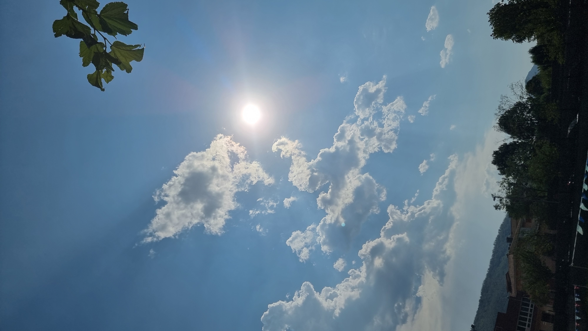 A vertical, outdoor, full-shot photograph captures a bright, sunny day with a clear blue sky dominating the upper portion. The sun is prominently positioned to the right, emitting strong rays that create a lens flare effect. Scattered white cumulus clouds are visible across the sky, some appearing fluffy and bright, while others are darker and more defined. In the upper right corner, a branch with green leaves extends into the frame. Below the sky, a distant, hazy mountain range can be seen. In the foreground, the roofs of buildings, some with red tiles, are partially obscured by a line of green trees. The bottom of the image reveals a dark foreground area, possibly a fence or netting, with faint outlines of decorative flags or banners. The scene appears to be set in Sant Joan Les Fonts, Spain, given the architectural style of the buildings and the surrounding landscape. There are no visible people or active events depicted in the photograph.