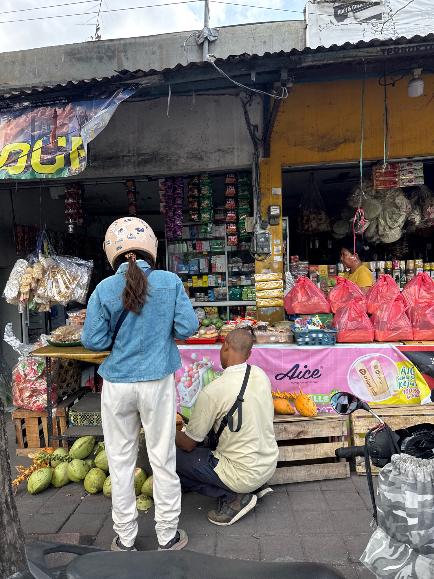 A medium shot shows a street-level view of a small shop in an urban environment. A woman wearing a light blue denim jacket, white pants, and a beige helmet with various stickers on it stands facing away from the camera. Her brown hair is tied in a ponytail. To her right, a man wearing a tan t-shirt and dark pants is crouched down. He has a black bag strap across his chest.
In front of the shop, a wooden table is laden with goods, including bags of snacks and what appear to be crackers. To the left of the table, several green coconuts and some yellow fruit are piled on the ground. Behind the table, shelves are filled with various packaged goods, and a pink banner with text and images is displayed. A woman in a yellow shirt is visible inside the shop, behind the counter, looking towards the right.
The shop itself has a yellow facade and an corrugated metal roof. Wires and conduits are visible along the exterior. The pavement in front of the shop is made of gray concrete slabs. A portion of a black motorcycle is visible in the foreground, on the right side of the image. The sky above is overcast.