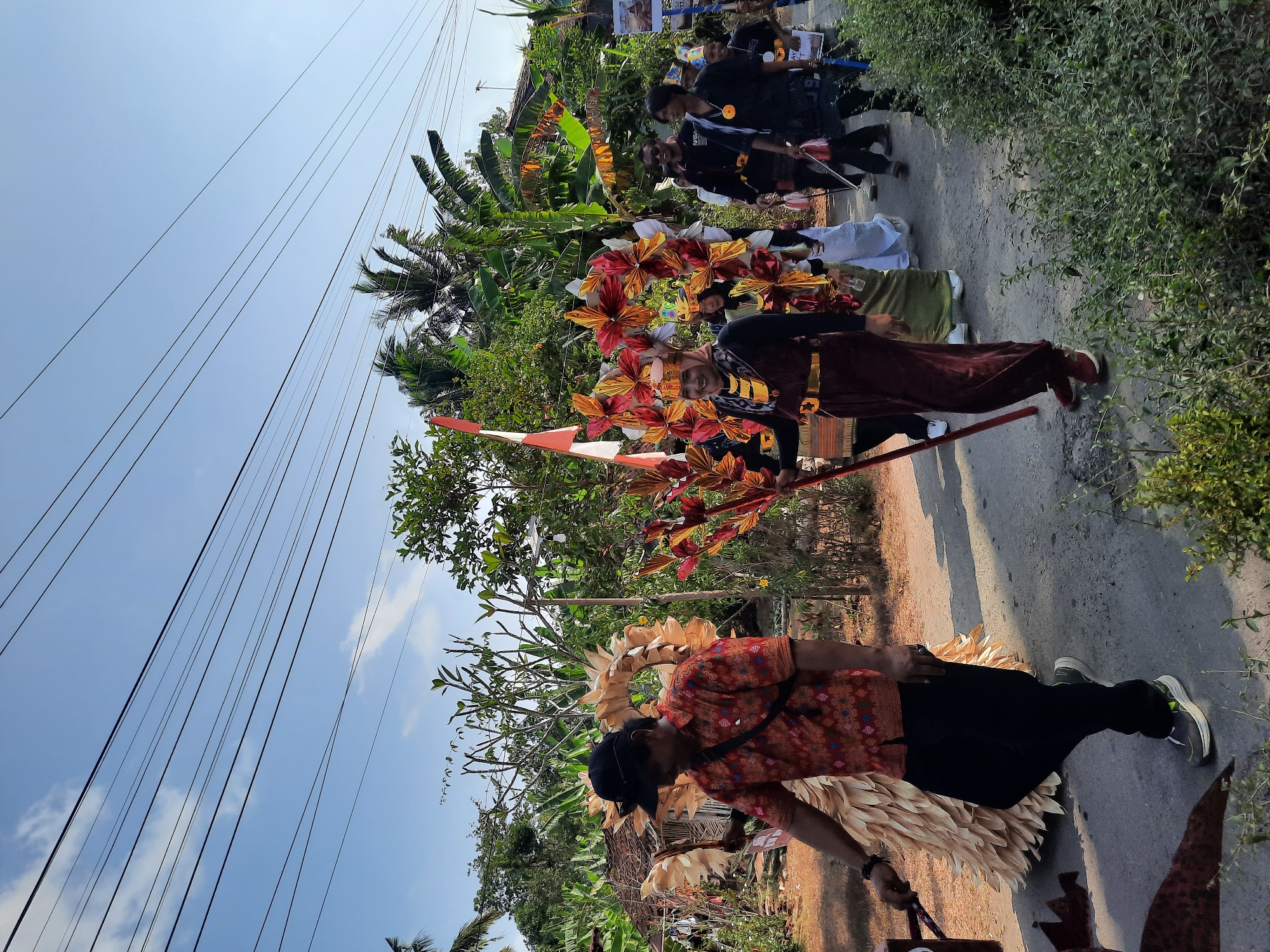 A group of people are marching in a parade on a sunny day in Andong, Indonesia. The parade participants are dressed in elaborate costumes adorned with colorful flowers and natural materials. In the foreground, a man in an orange shirt and black pants walks alongside a large, straw-like costume. Behind him, several individuals are walking, some holding staffs decorated with flowers. The background features lush green trees, banana plants, and a few buildings. Power lines are visible across the blue sky. The road they are walking on is paved with asphalt.