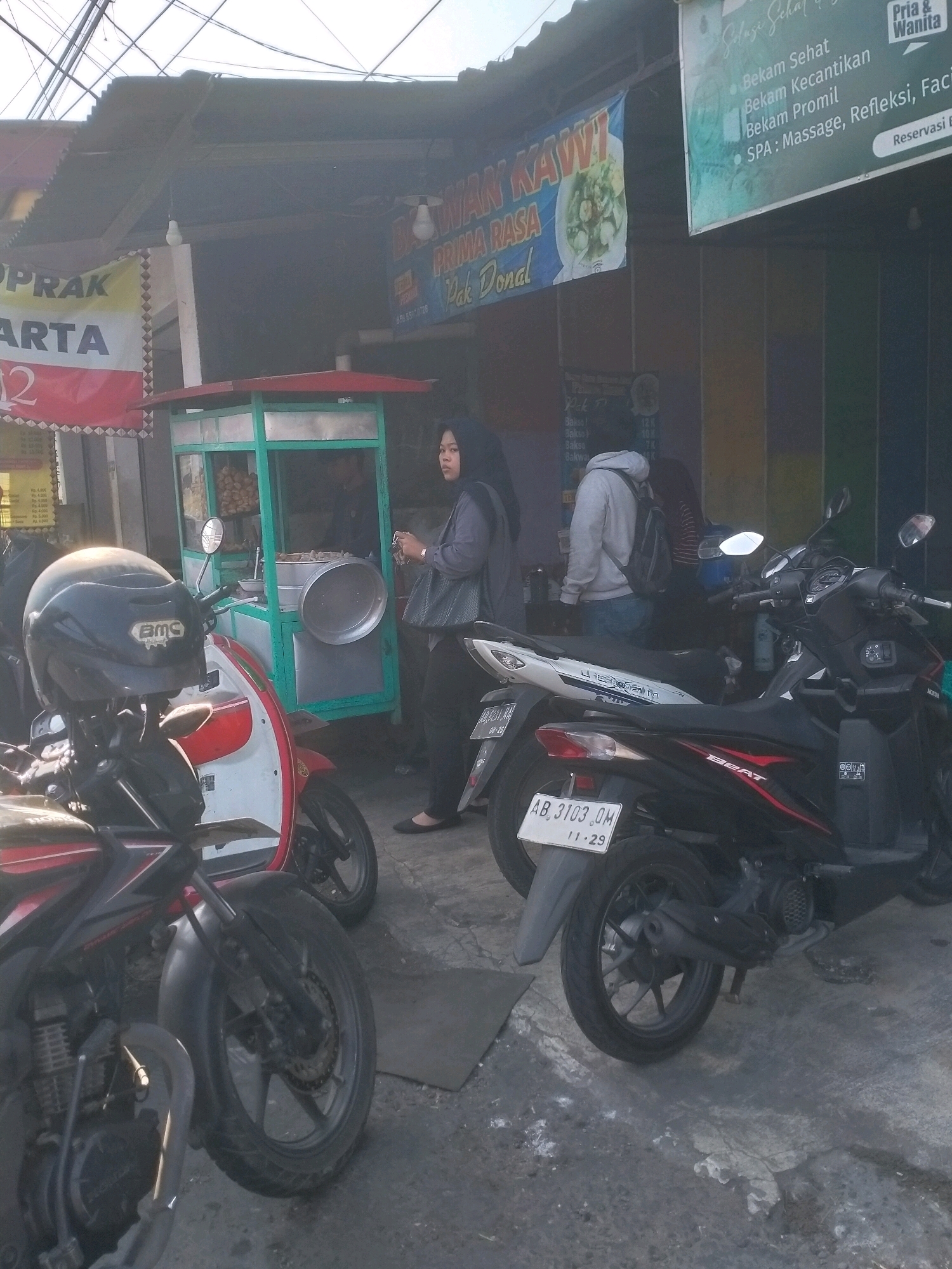 The image depicts a street scene in Kasihan, Indonesia, likely in the late afternoon judging by the lighting. In the foreground, two motorcycles are parked. The one on the left is a dark-colored sportbike with a black helmet on its handlebars. To its right is a red and white scooter. Behind the motorcycles, a food stall with a green frame and a glass display case is set up. Several people are visible near the stall. A woman in a gray hijab and dark clothing stands to the right of the stall, looking towards the camera. Behind her, a person wearing a light gray hoodie and jeans stands with a backpack. A third person in a dark shirt is visible inside the stall. Two banners hang above the stall; one is red and white with text, and the other, further back, is blue with white text and an image of food. The ground is paved and appears somewhat worn. The background shows the facade of buildings with electrical wires overhead. A sign on the right side of the image is green with white text listing spa services.