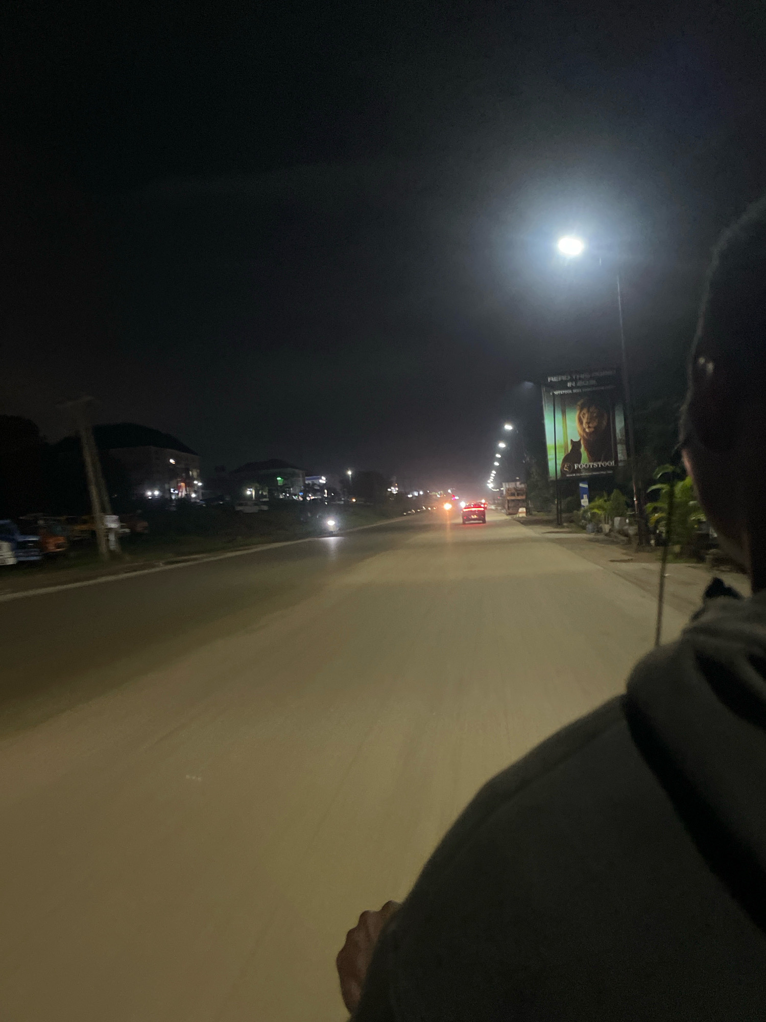 An unidentifiable person, wearing a dark hooded jacket, is viewed from behind, riding on a moving vehicle at night, likely a motorcycle, as they look forward along a paved road in Abuja, Nigeria. The road is illuminated by streetlights and the headlights of distant cars. On the right side of the road, a large billboard features an image of a lion and a cat, with the text "READ THIS IMPORTANT IN NIGERIA." and the brand name "FOOTSTOOL." Further down the road, buildings and more streetlights are visible. The sky is dark and overcast, suggesting it is nighttime in the city.